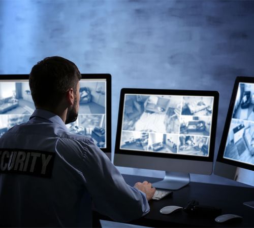 A security guard is sitting in front of three computer monitors