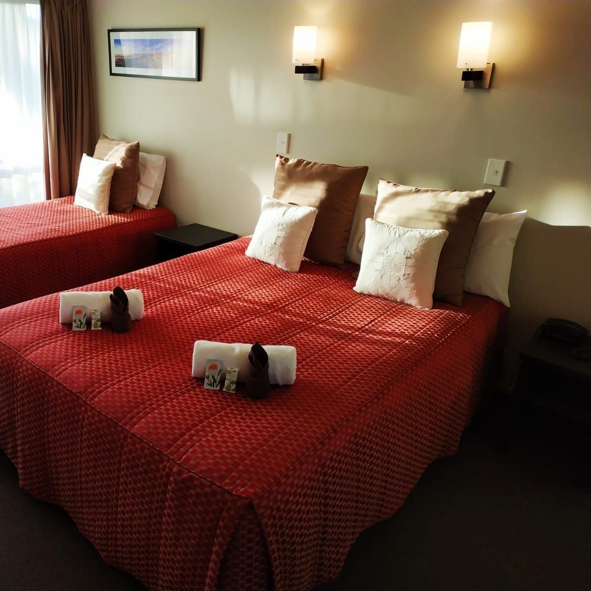 Two beds in a hotel room with red bedspreads, pillows, and side tables.