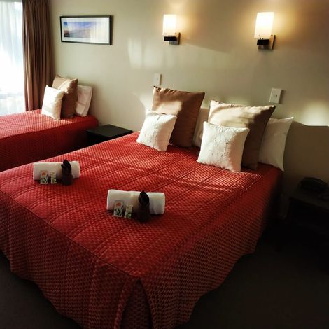 Two beds in a hotel room with red bedspreads, pillows, and side tables.