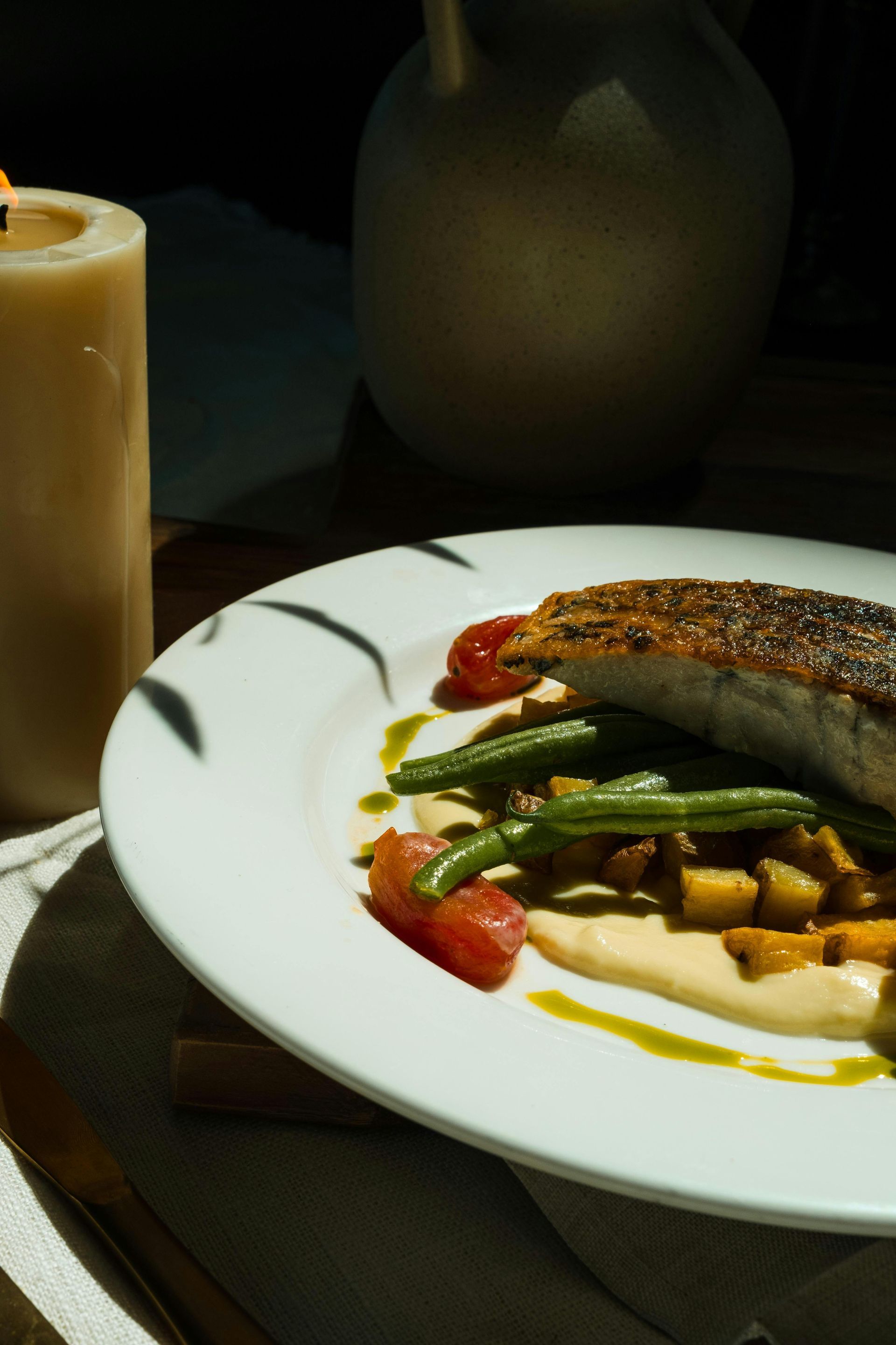 Plate of seared fish with vegetables and sauce, lit by candlelight.