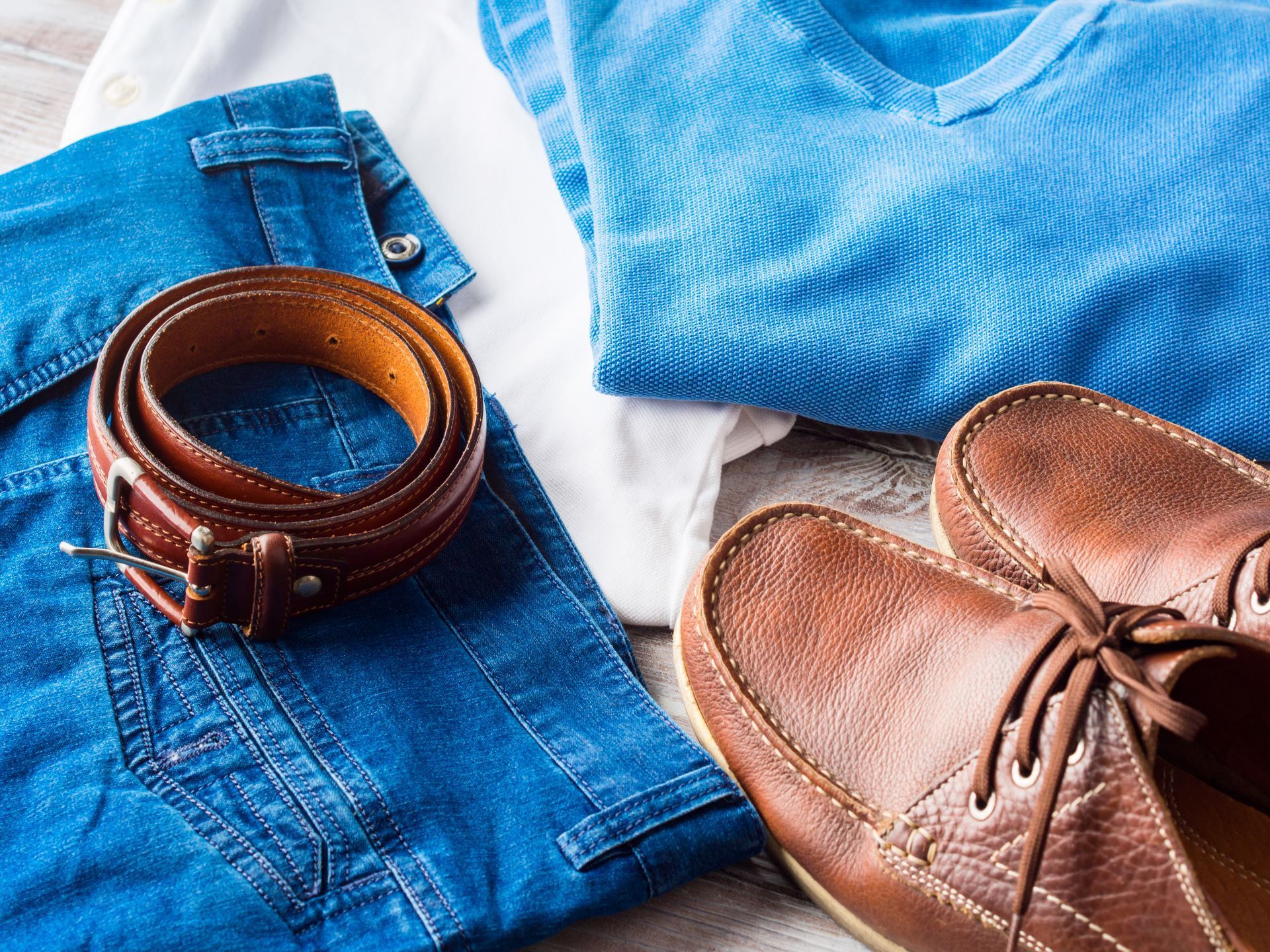 A pair of brown shoes , a belt , a shirt , jeans and a sweater are on a table.