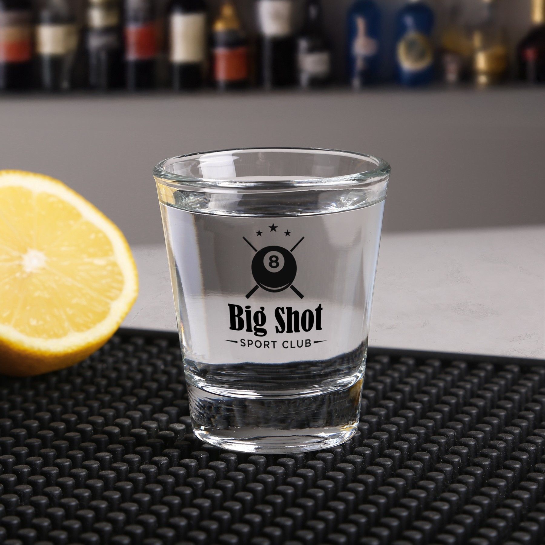 A shot glass with a logo on it is sitting on a bar mat next to a slice of lemon.