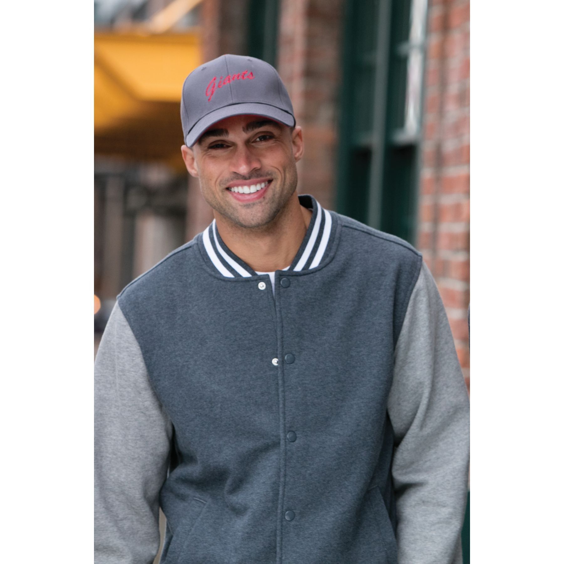 A man wearing a baseball cap and a varsity jacket is smiling.
