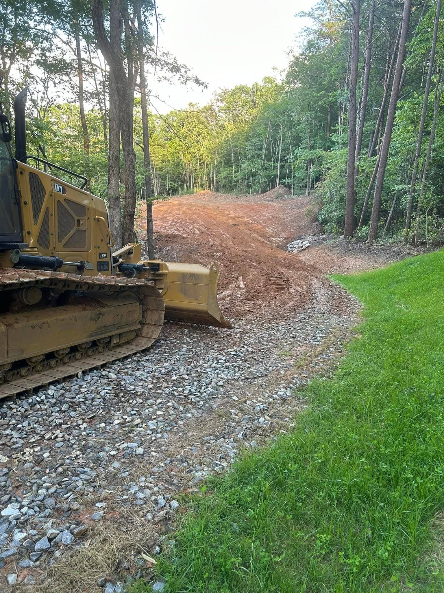 A yellow bulldozer clearing a dirt path through a wooded area next to a gravel driveway and green grass.