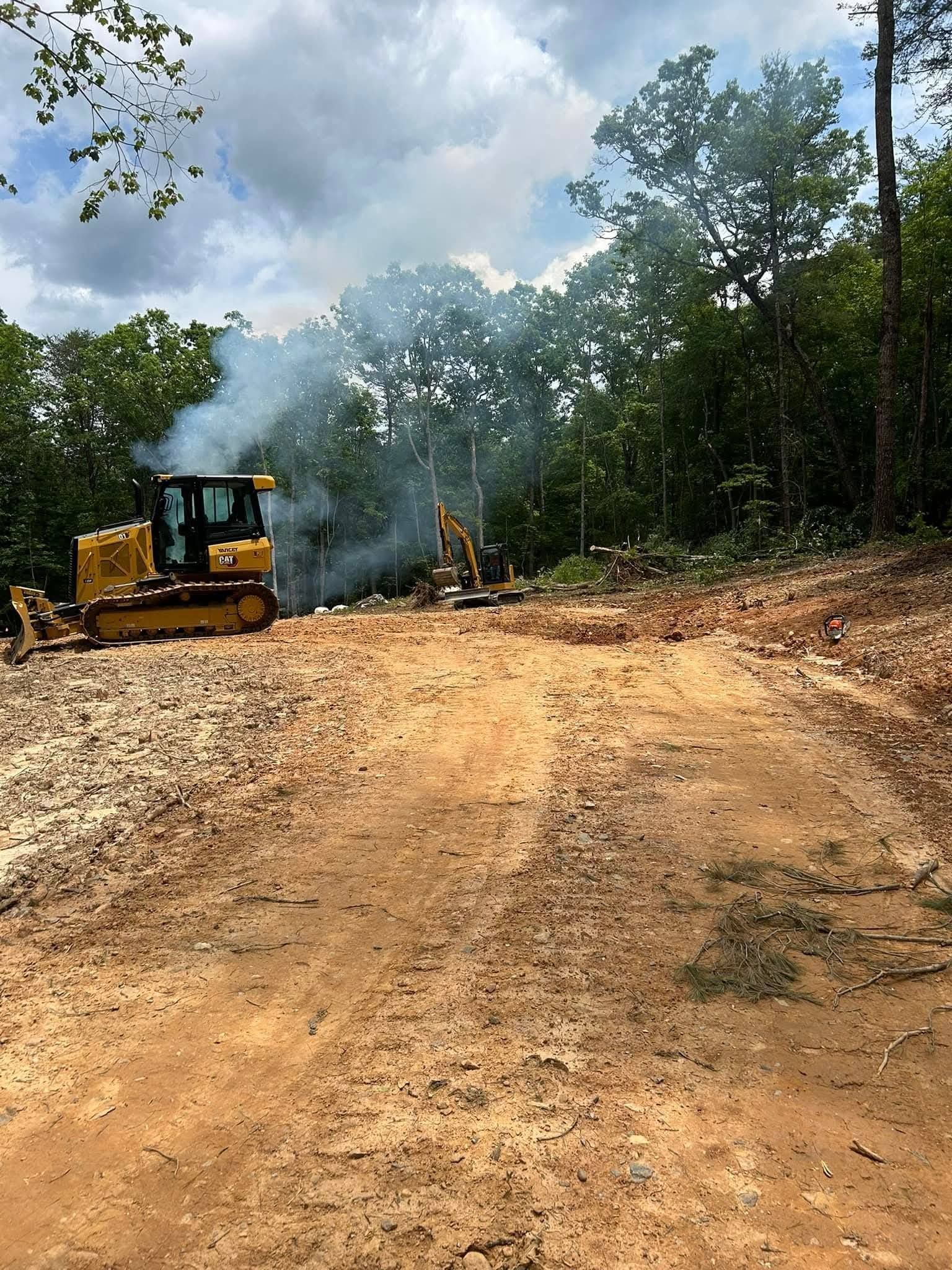 A yellow bulldozer sits on a cleared dirt lot with smoke rising from behind it near a treeline.