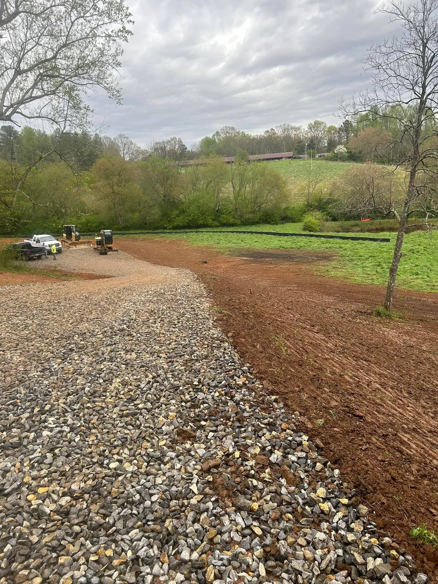 A gravel driveway bordered by brown mulch leads toward a green field and a line of trees under a cloudy sky.