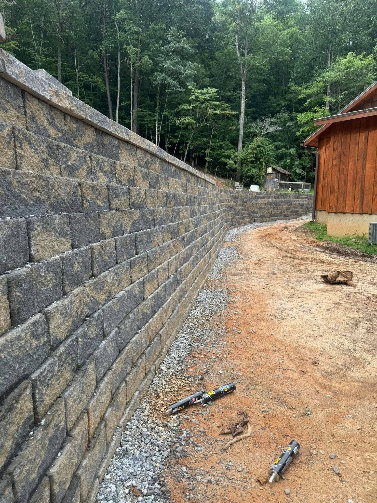 A tall, multi-colored stone retaining wall curves along a dirt path next to a wooden shed near a forest.