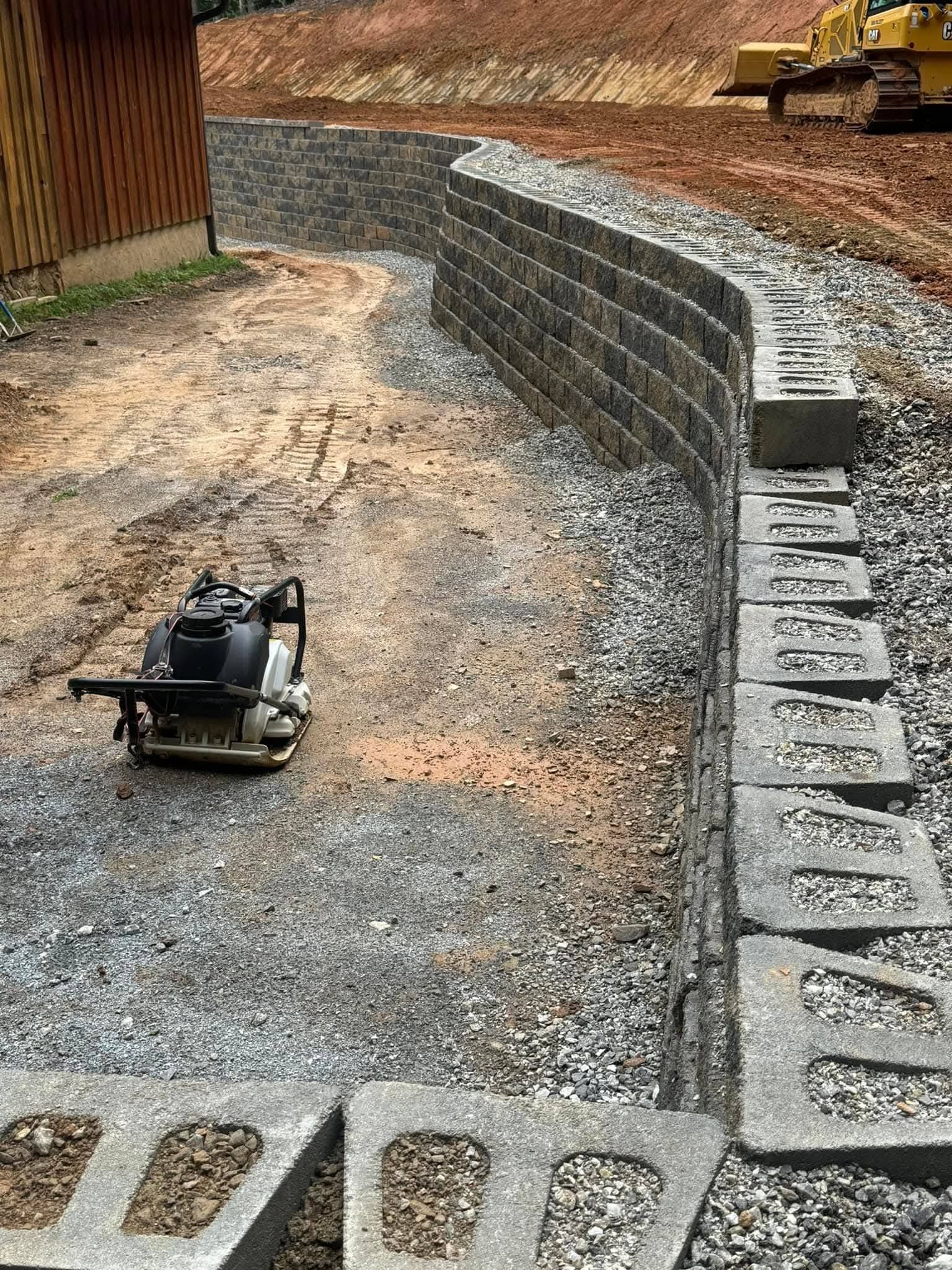 A plate compactor sits on a gravel base next to a curved retaining wall built from interlocking gray concrete blocks.