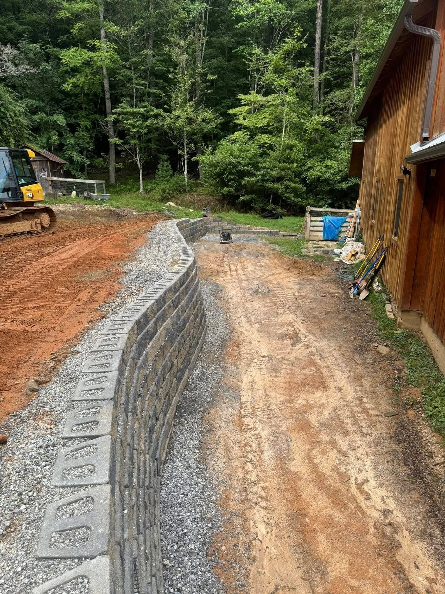 A curved stone retaining wall under construction next to a dirt path and a wooden building, with forest in the background.