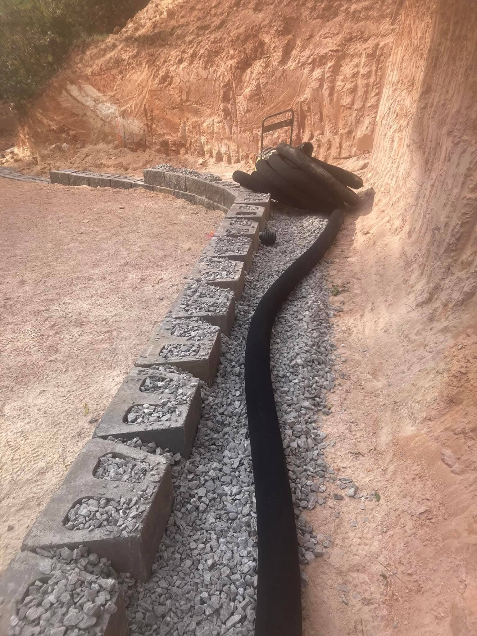 A drainage pipe lies along a gravel trench behind a curved concrete block retaining wall next to a dirt slope.