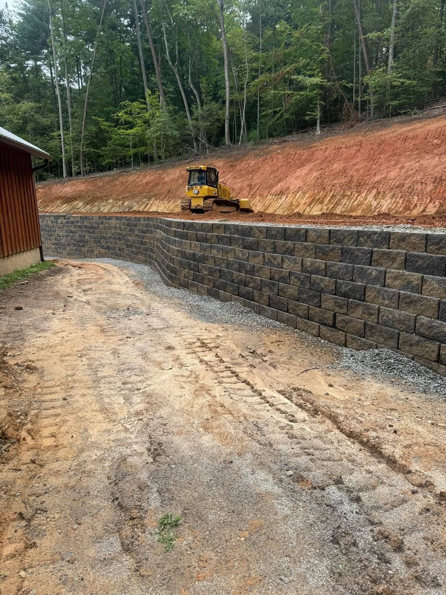 A yellow bulldozer sits atop a tiered retaining wall made of dark stone blocks bordering a dirt construction path.