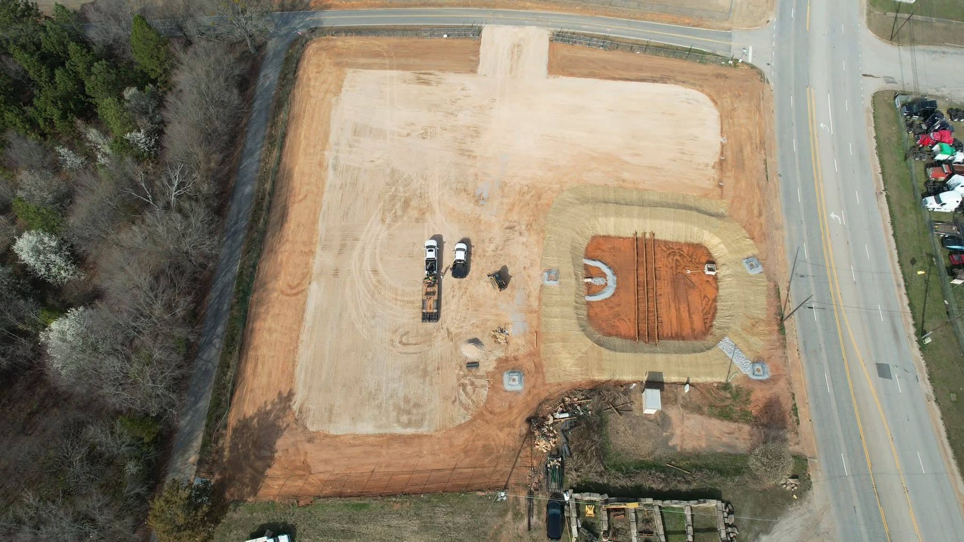 Aerial view of a cleared construction site with a foundation pit and two parked vehicles near a road and wooded area.