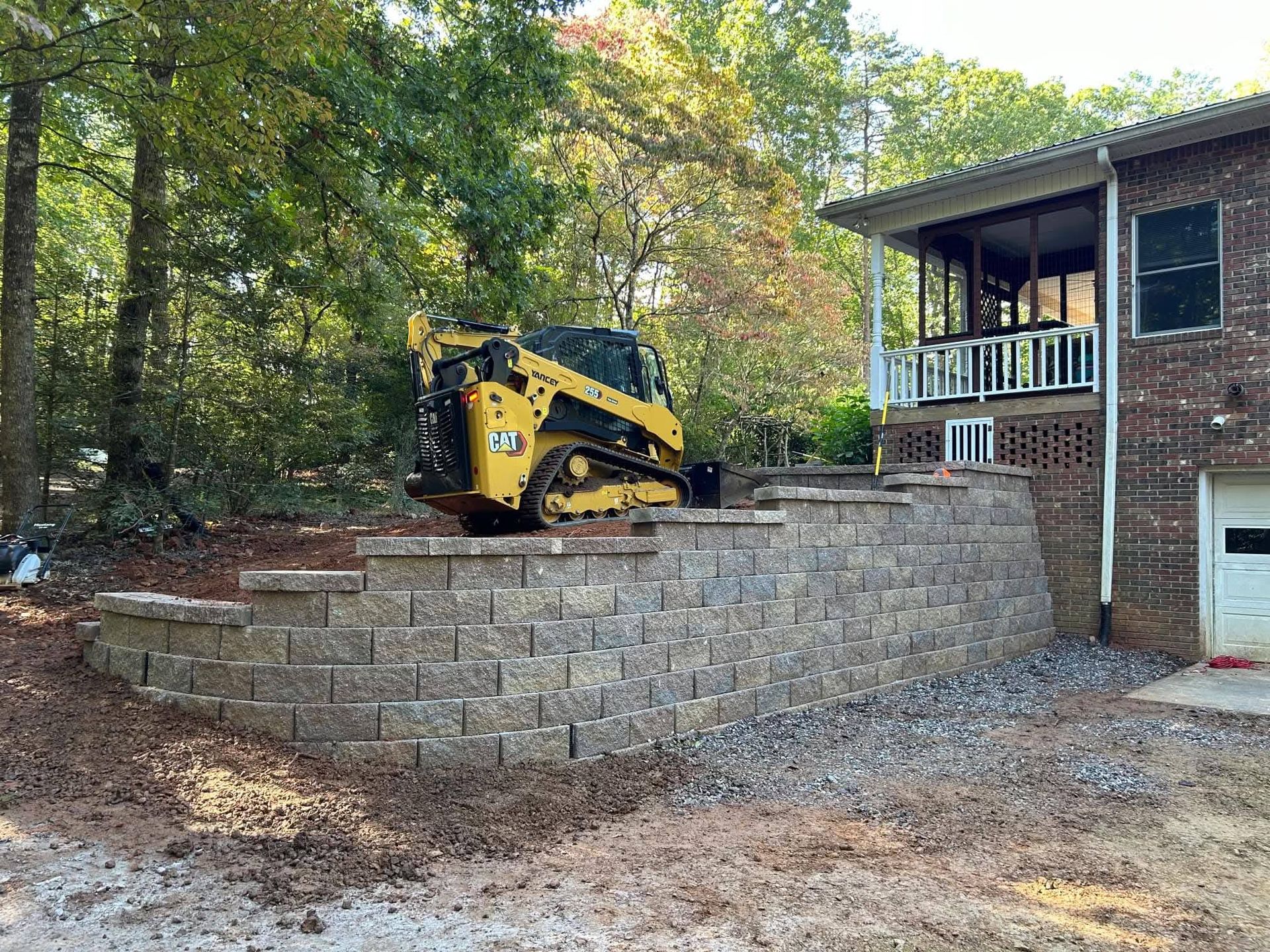 A yellow skid steer loader sits atop a newly constructed, multi-tiered stone retaining wall next to a brick house.