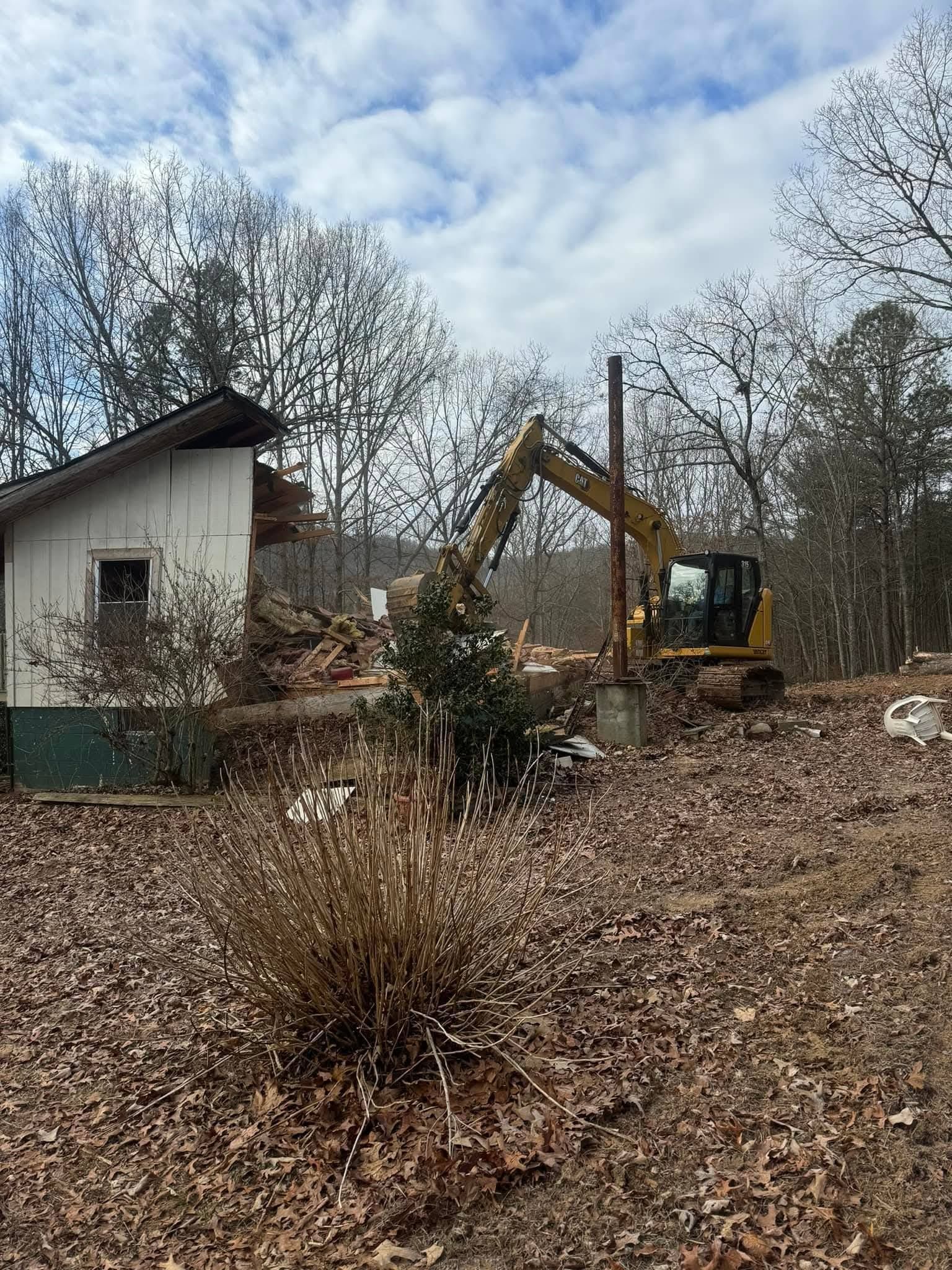 A yellow excavator demolishes a small white house in a wooded area covered with fallen brown leaves.