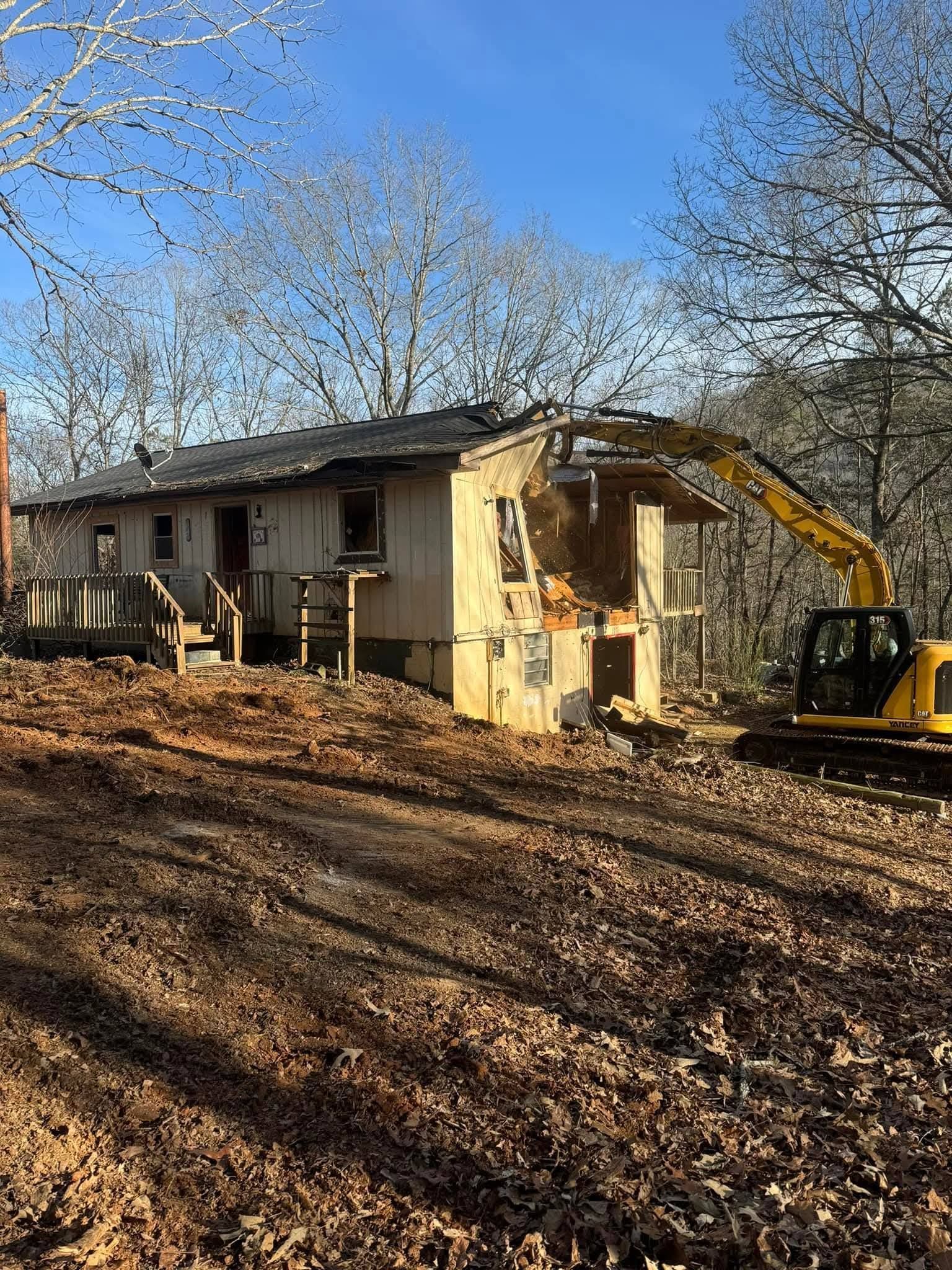 A yellow excavator tears into the side of a small, light-colored house located on a wooded, leaf-covered hill.