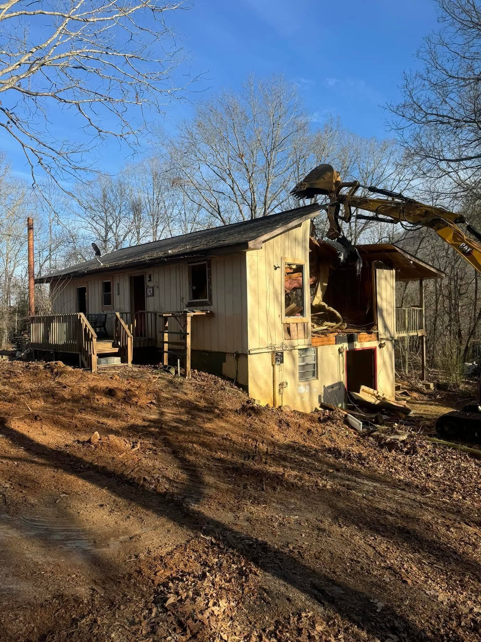 An excavator demolishes a wood-sided house in a wooded area under a clear blue sky.