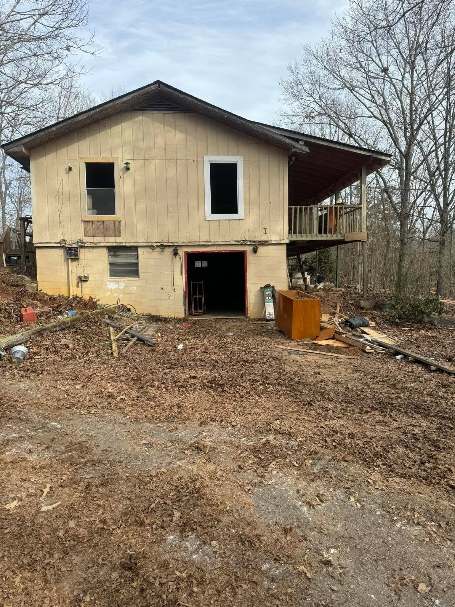A two-story beige house with an open garage, a deck on the right side, and a yard covered in fallen brown leaves.