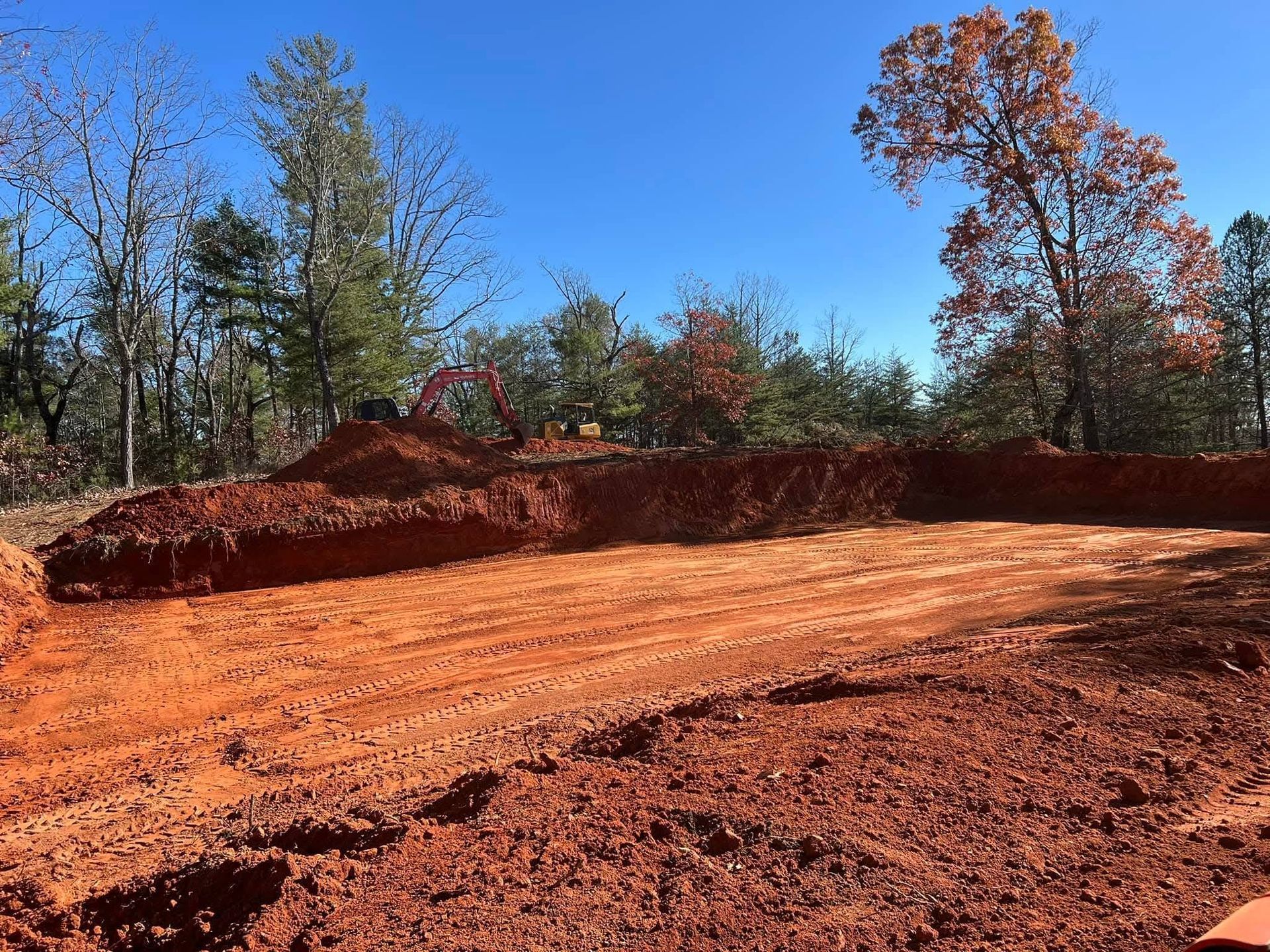 A construction site with a deep excavated pit of red clay soil surrounded by a forest under a clear blue sky.