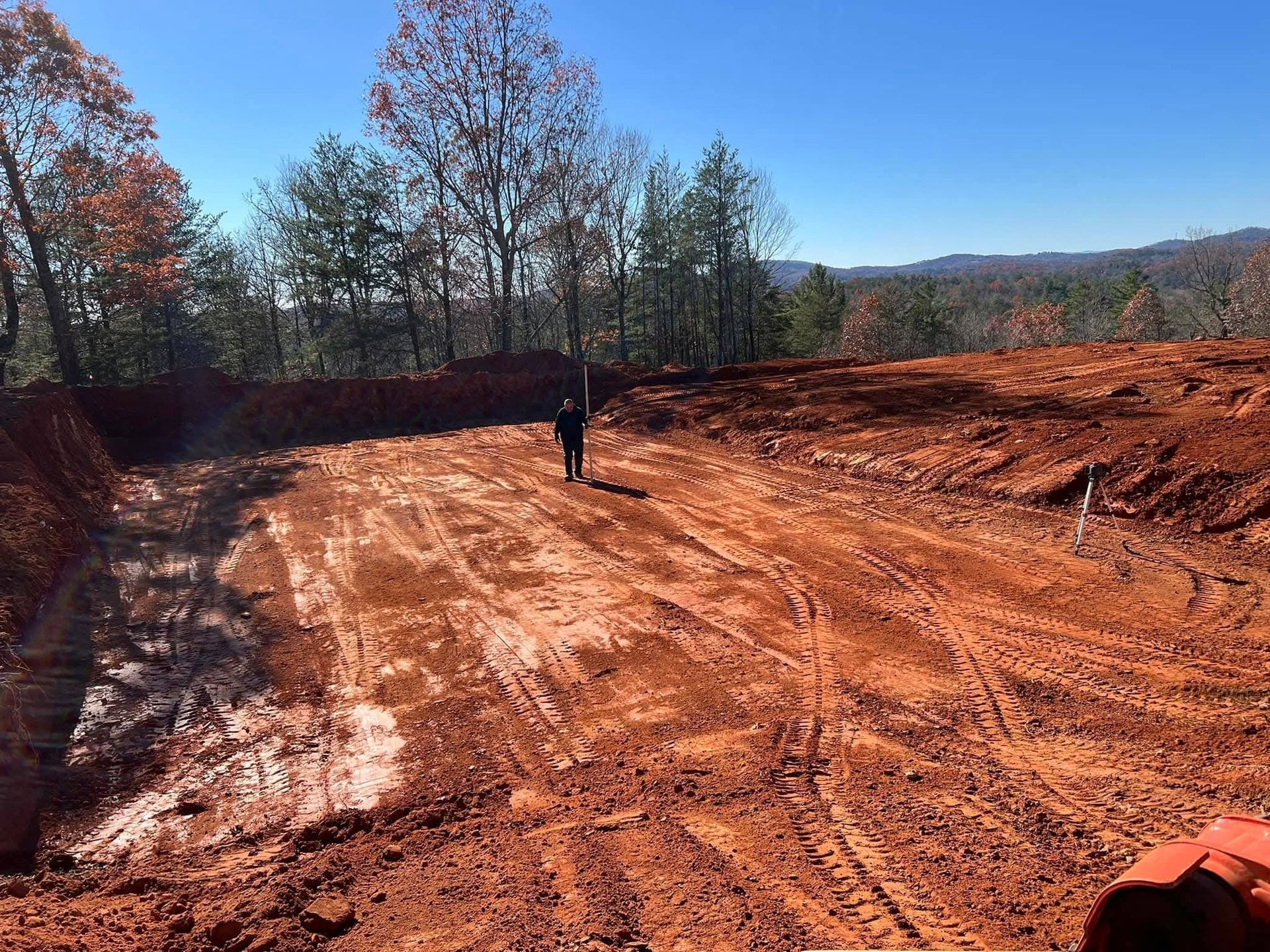 A person stands in a large, cleared red-dirt lot prepared for construction, bordered by trees under a clear blue sky.