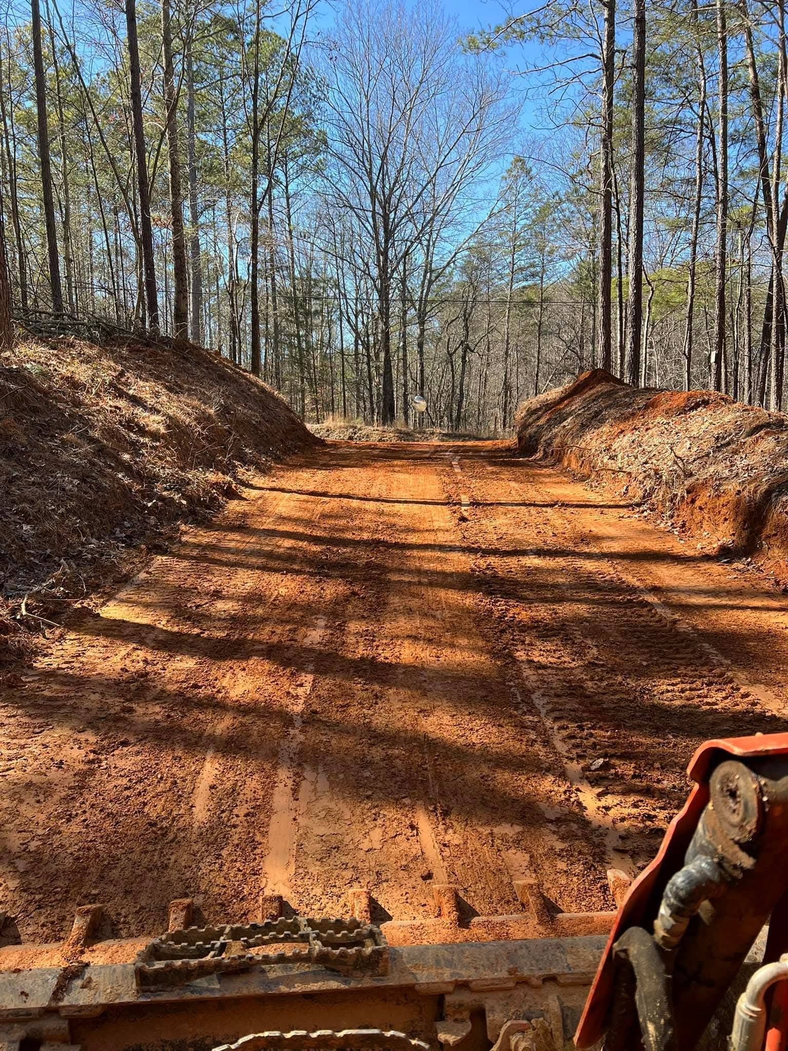 A view from the cab of heavy machinery overlooking a cleared, red dirt path carved through a pine forest.