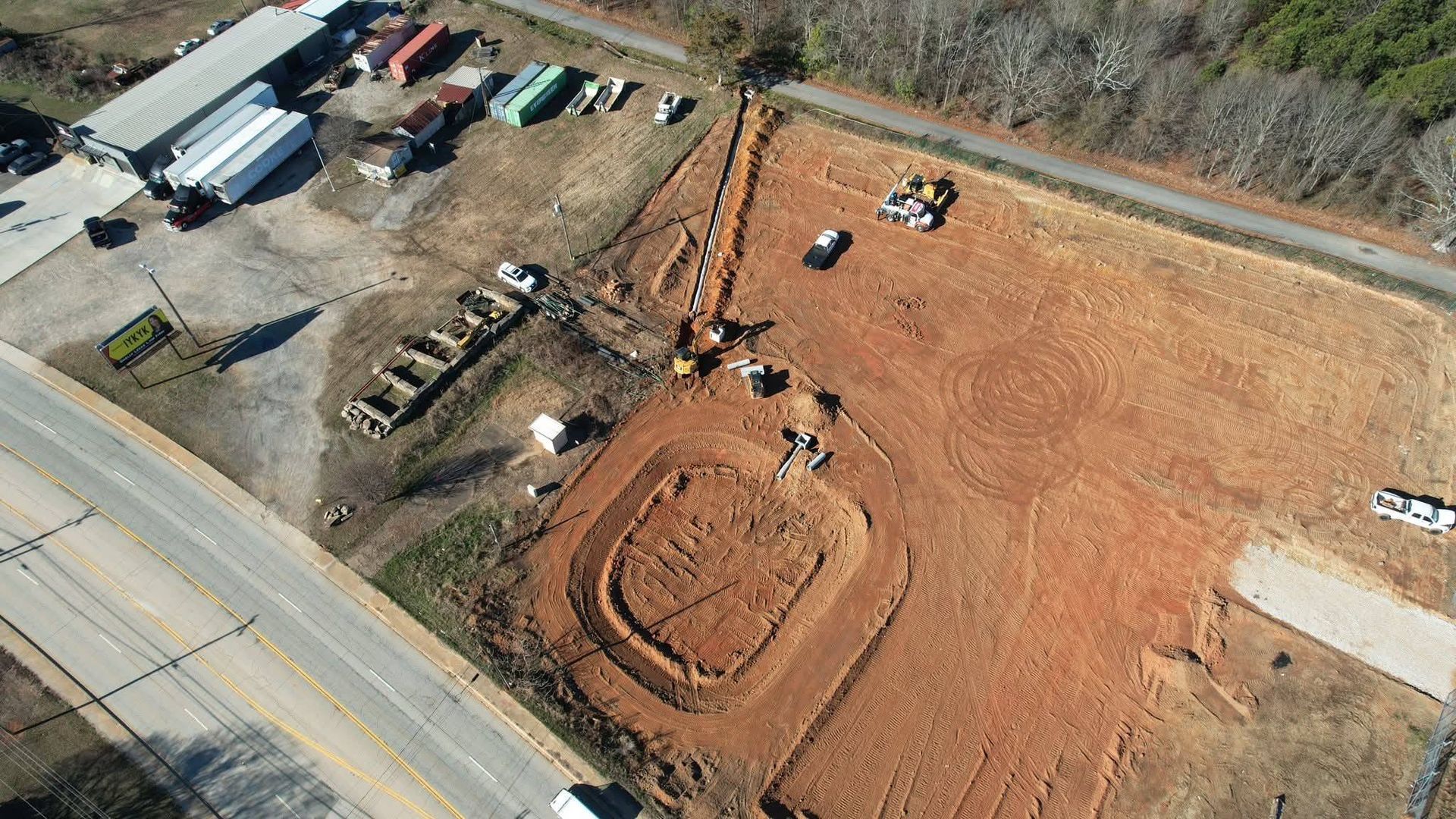 An aerial view of a construction site with an excavated rectangular pit, trenches, dirt mounds, and parked vehicles.