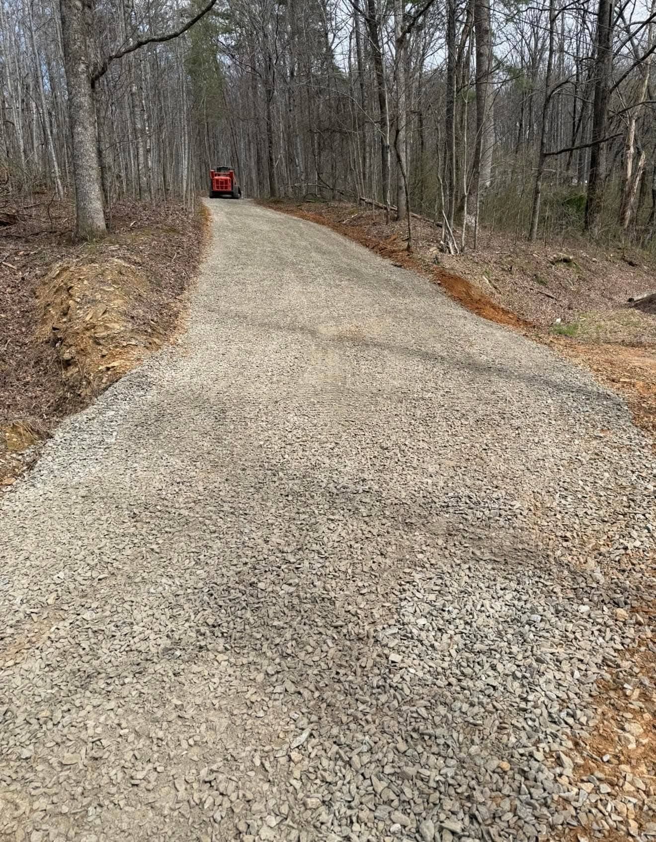 A gravel driveway winds through a wooded area with a small red vehicle parked in the distance.