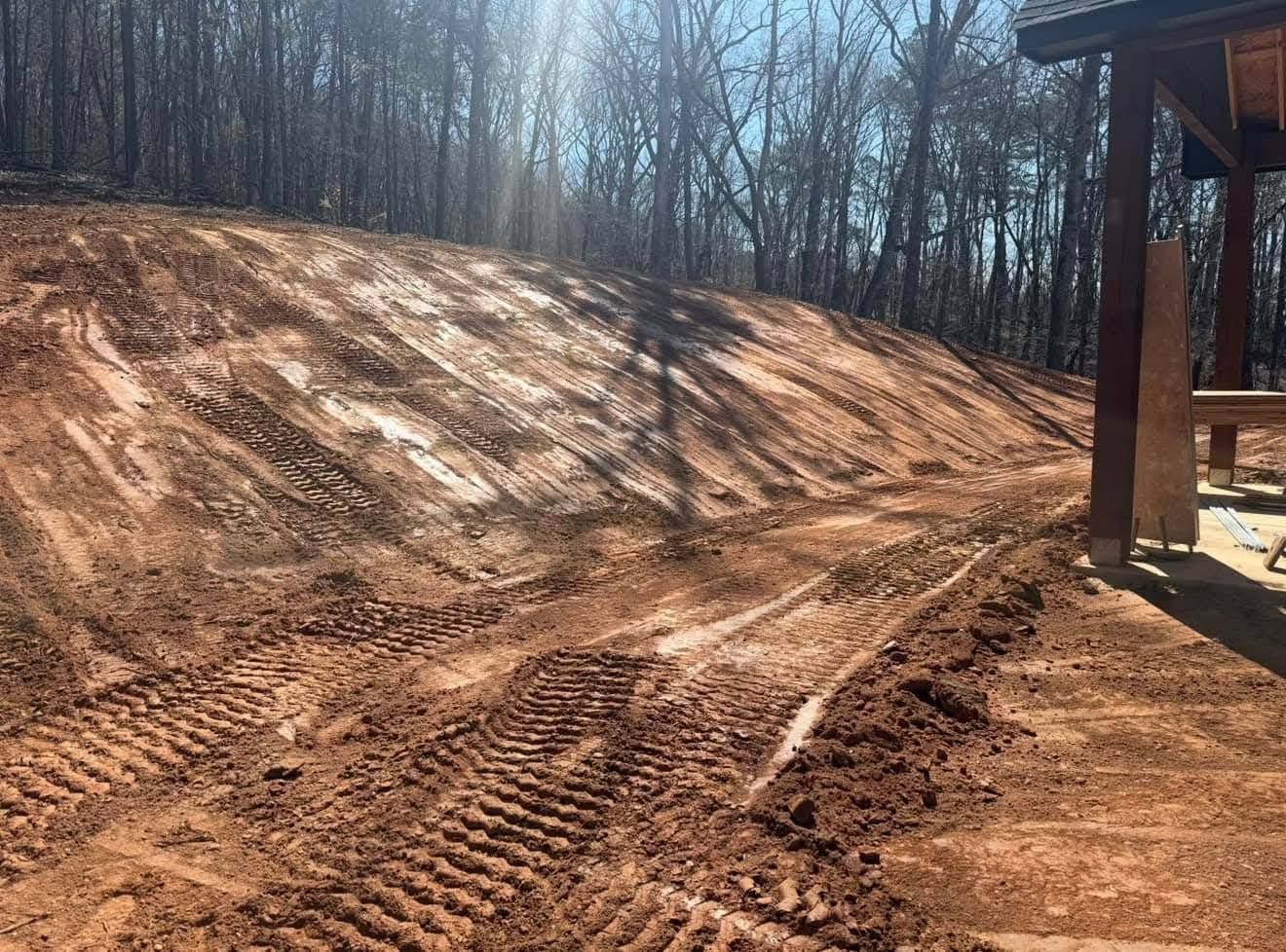 A wide, freshly graded earthen slope with prominent heavy equipment tracks, next to a wooden structure near a forest.