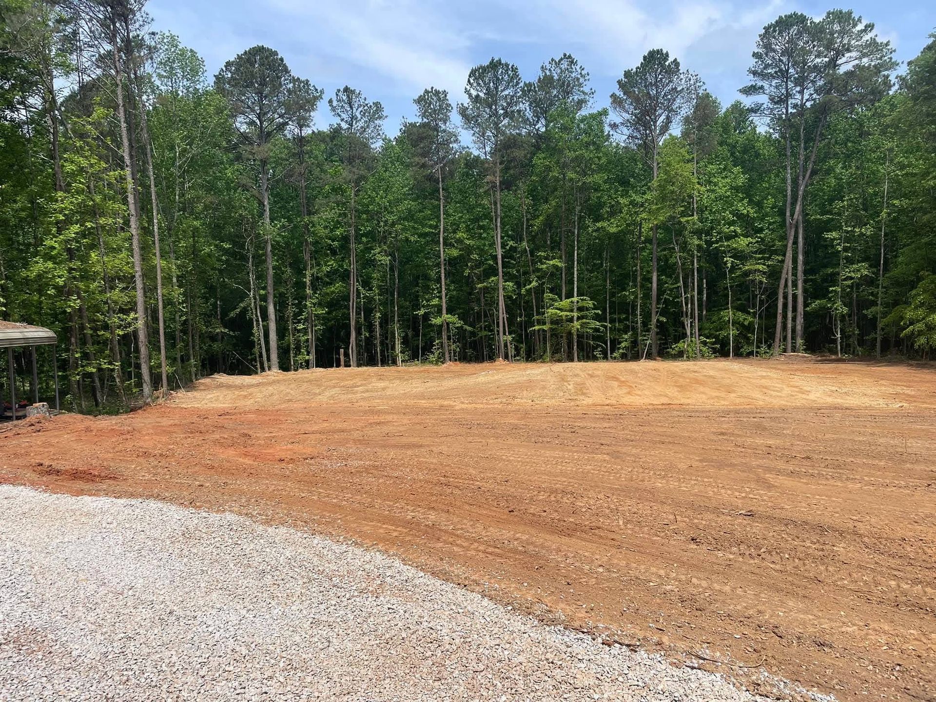 A gravel driveway borders a large, cleared patch of red dirt and soil in front of a dense forest of tall green trees.
