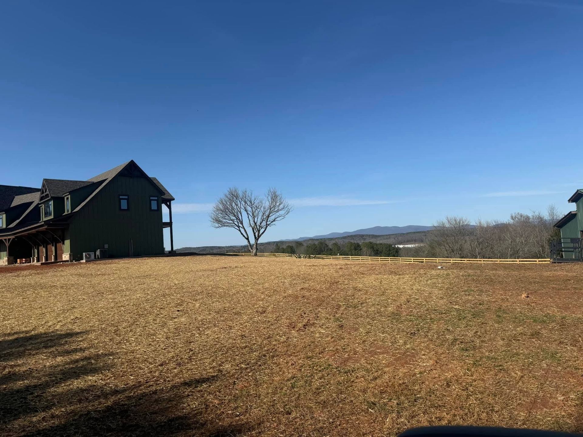A sprawling brown field under a clear blue sky, featuring a large house on the left and a lone tree in the distance.