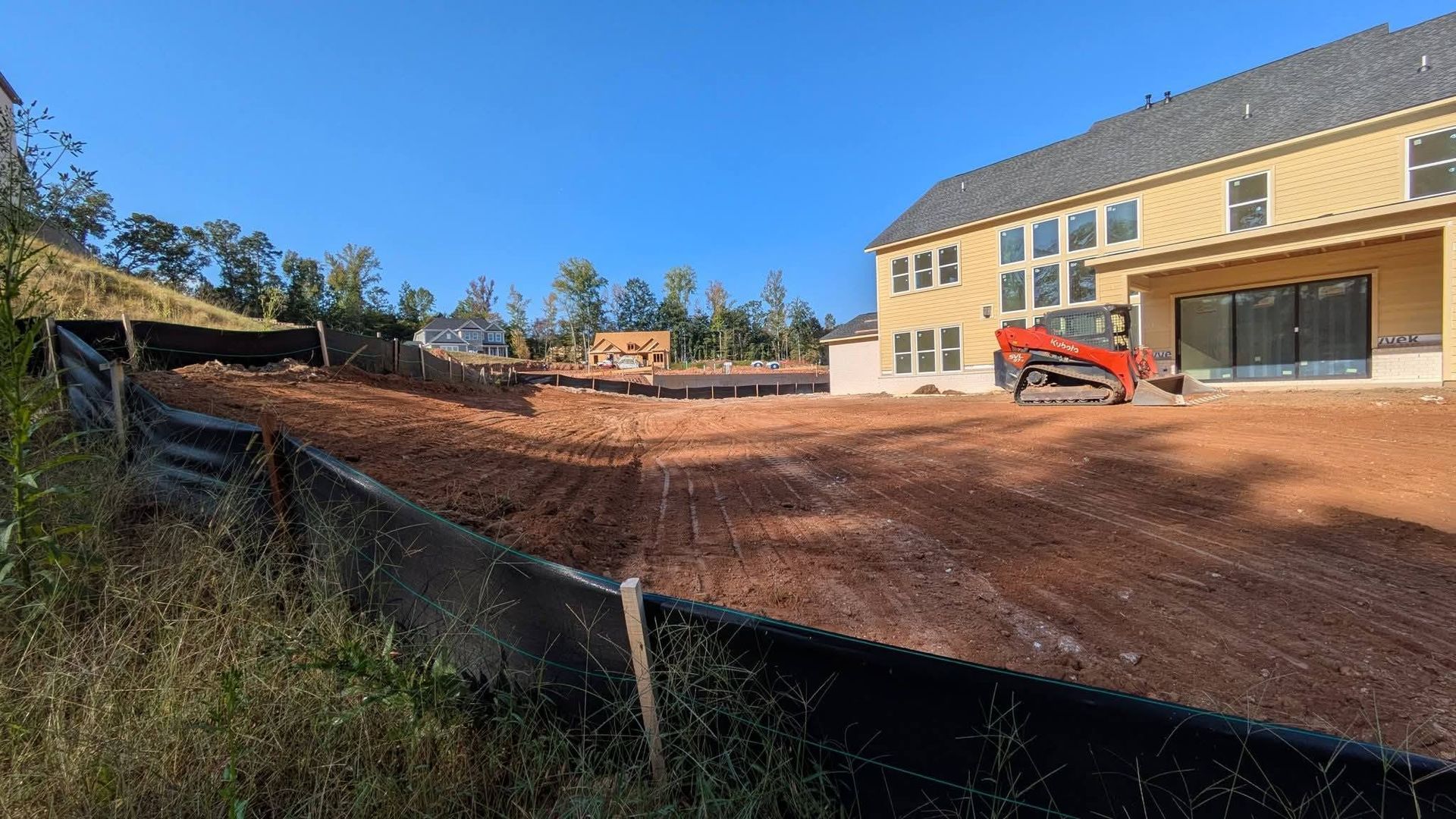 A newly graded, red-dirt construction site next to a large yellow house, bordered by a black silt fence under a blue sky.