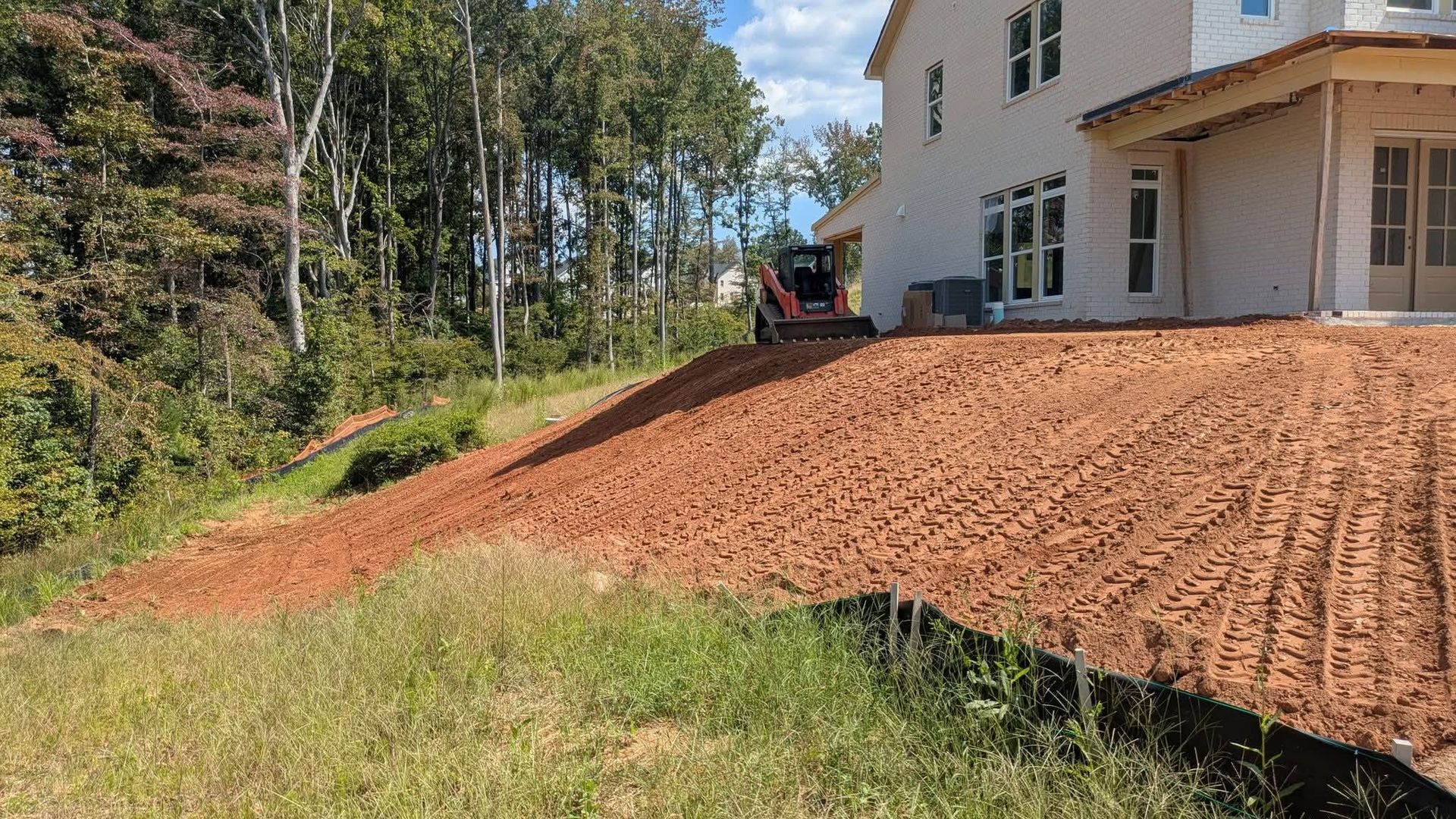 A construction site showing a newly graded red clay slope beside a house, with a small skid-steer loader on the hill.
