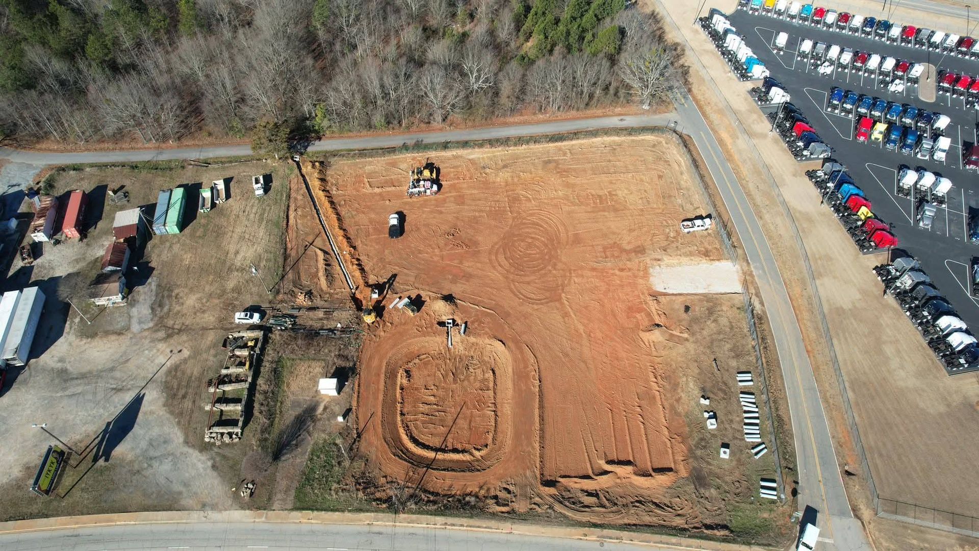 Aerial view of a cleared construction site with exposed red soil, trenches, and nearby parking lots with many vehicles.