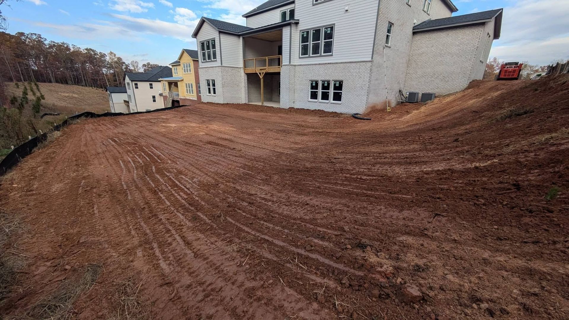 A new, unfinished house sits on a large, graded lot with reddish-brown soil under a partly cloudy sky.
