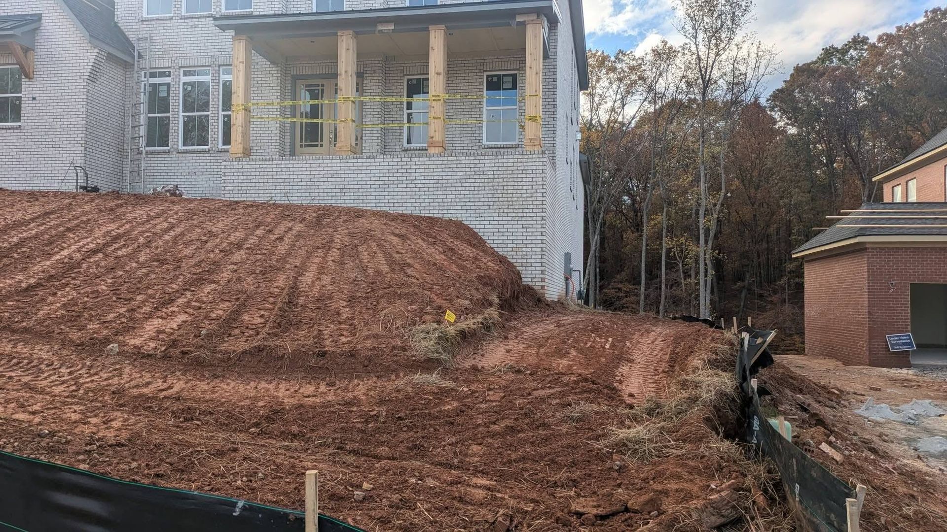 A two-story house under construction with a large, freshly graded dirt slope in the foreground and a wooded area behind.