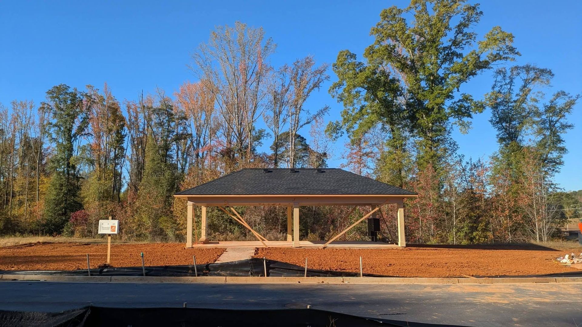 A small open-air wooden pavilion sits on a clearing of brown dirt in front of a line of trees under a clear blue sky.