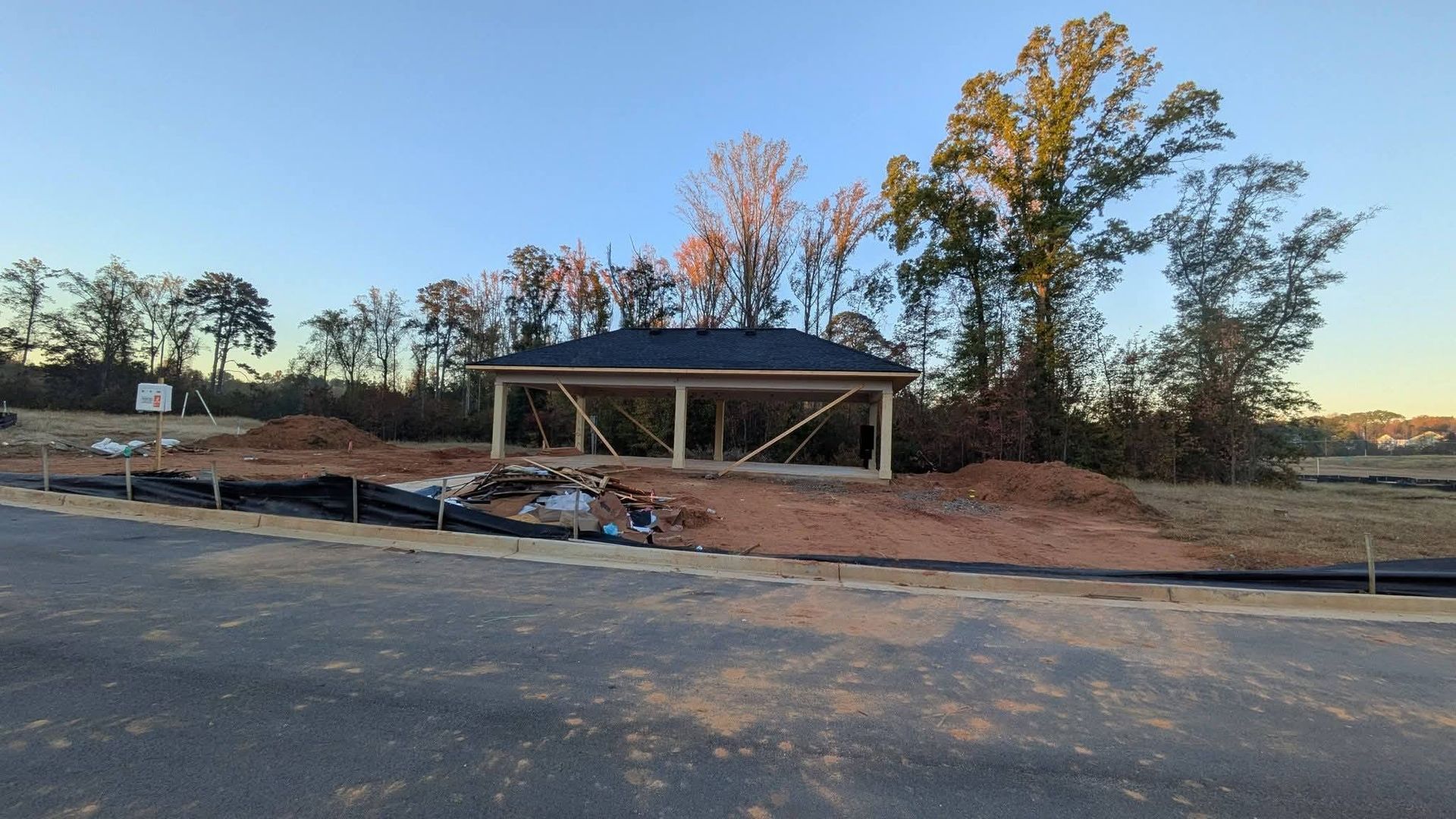 A wooden pavilion under construction at sunset, situated on a dirt lot next to a newly paved road.
