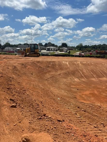 A yellow bulldozer sits on a leveled, red-clay dirt construction site under a partly cloudy blue sky.