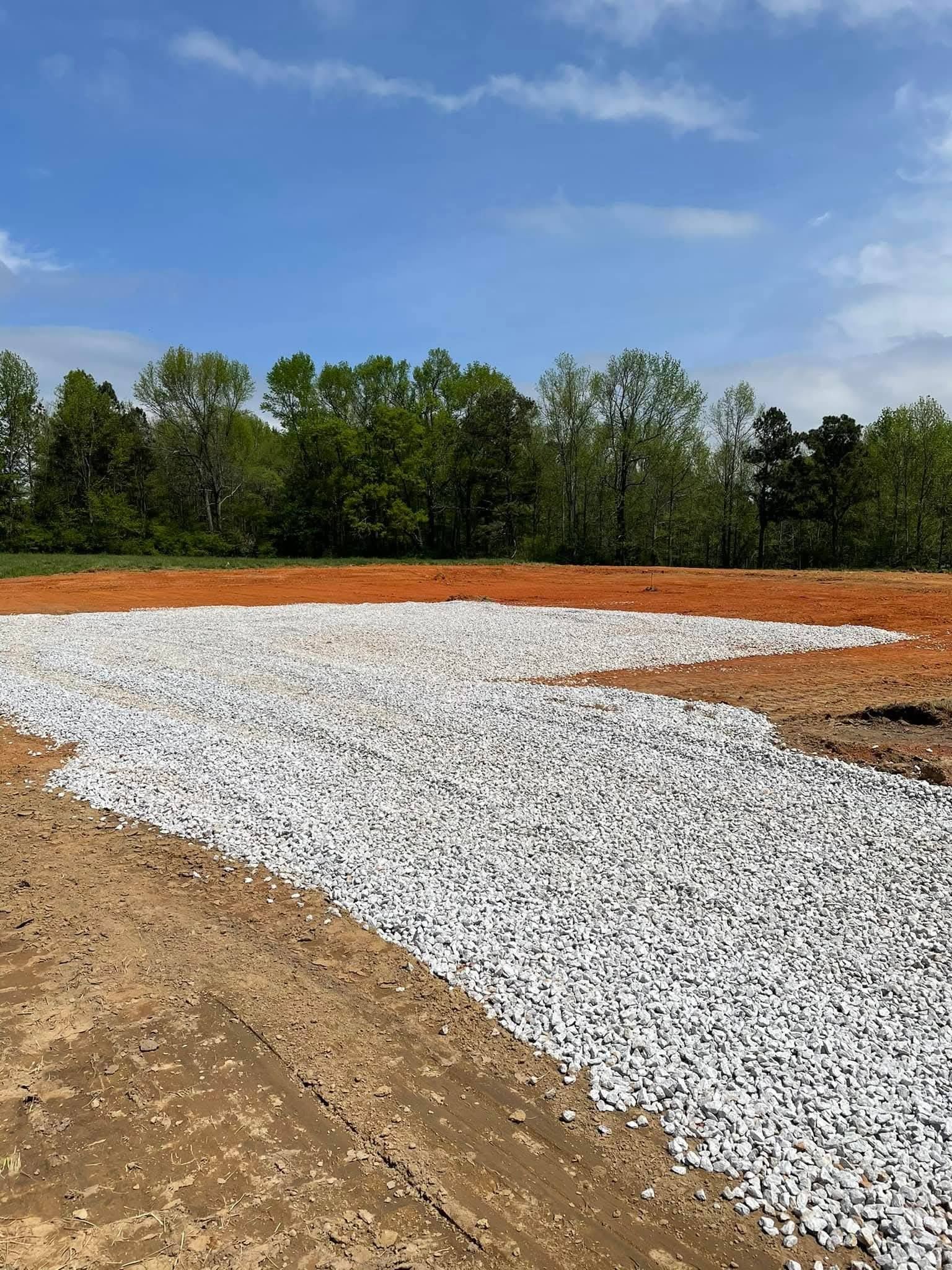 A patch of white gravel rests on cleared, red dirt ground beneath a blue sky with thin clouds and distant green trees.