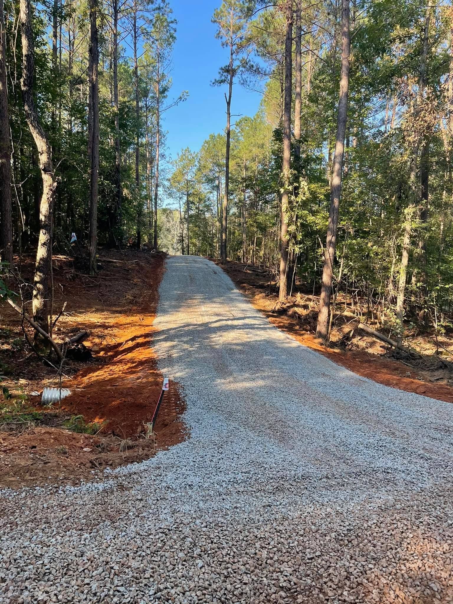 A gravel driveway winds uphill through a sunlit forest with red clay soil on the sides under a clear blue sky.