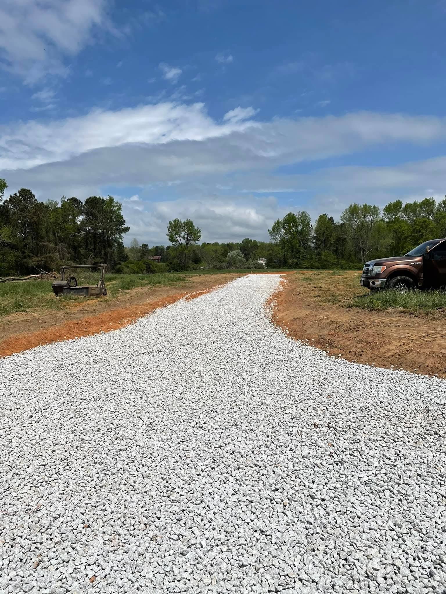 A gravel driveway stretches through a cleared dirt lot under a blue, cloudy sky, with a truck parked on the right.