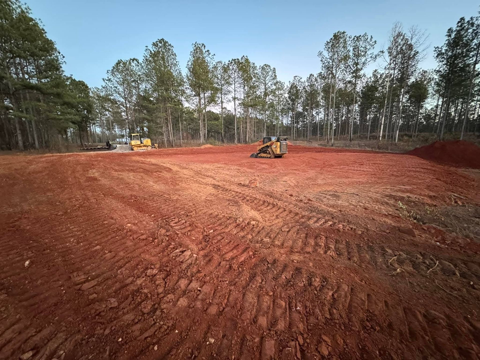 A construction site with reddish dirt, tire tracks, and heavy machinery parked near a forest under a clear sky.