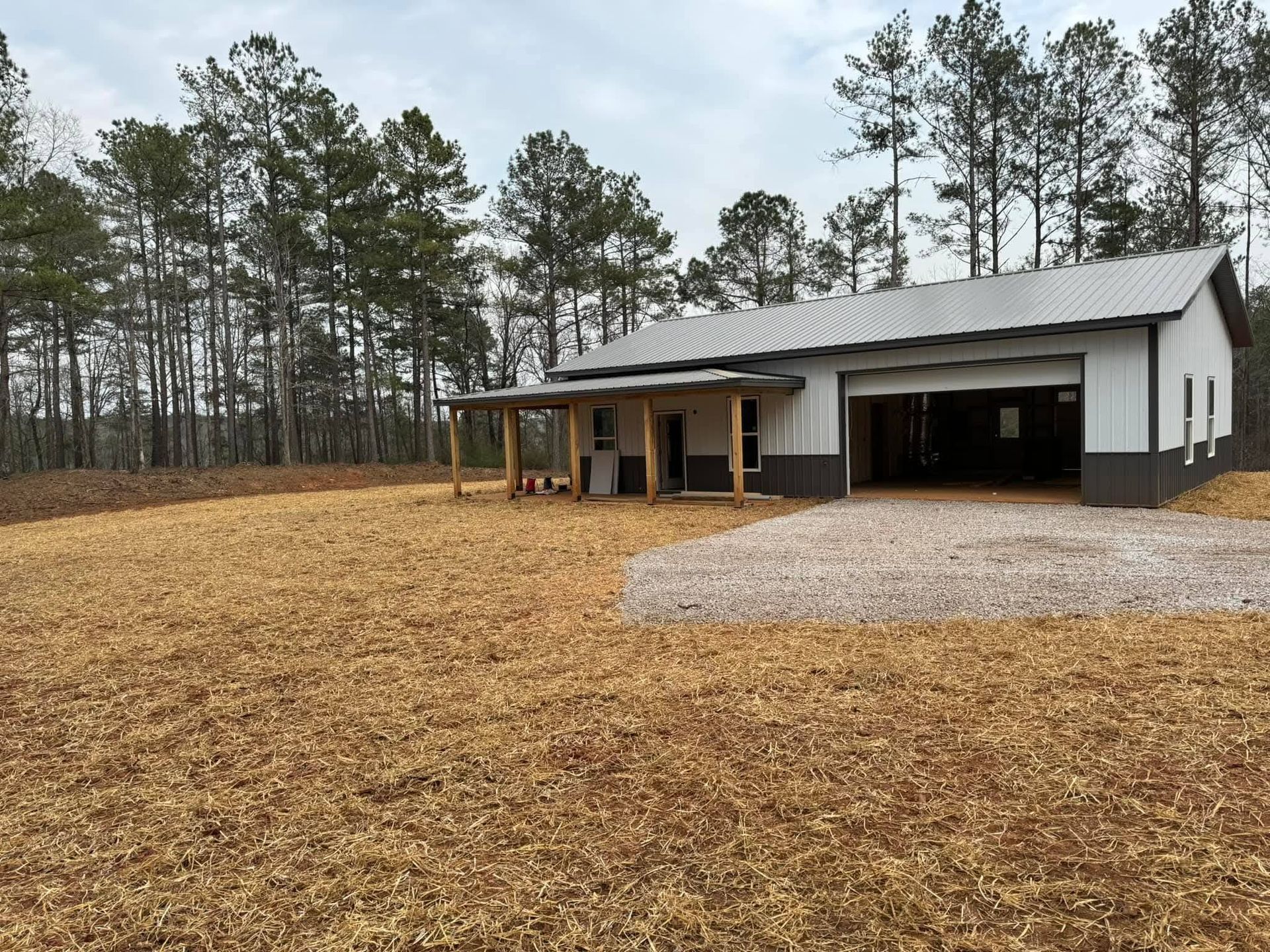 A modern barndominium-style house with a metal roof, grey walls, and a wooden porch set against a forest edge.