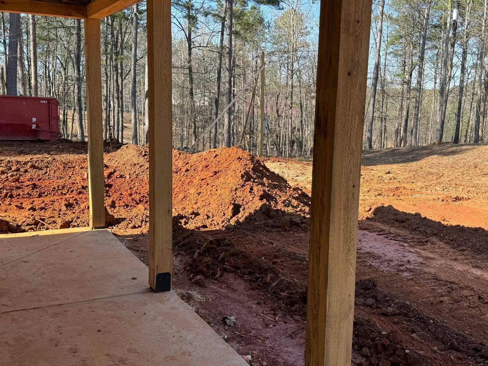 A wooden porch frame looks out over a construction site with piles of red clay earth and a forest in the background.