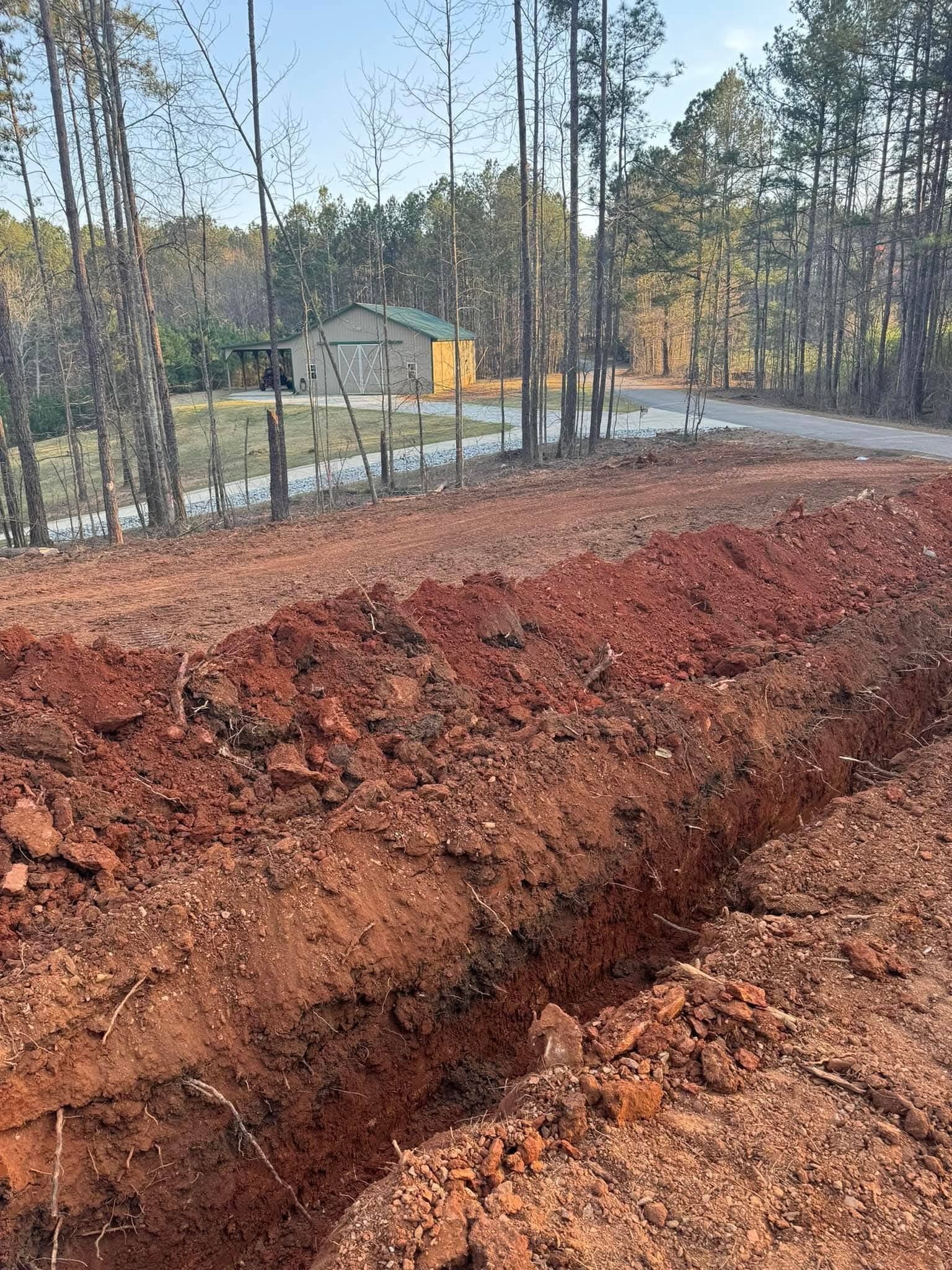 A long, freshly dug trench cuts through red dirt, with a house and trees visible in the background.