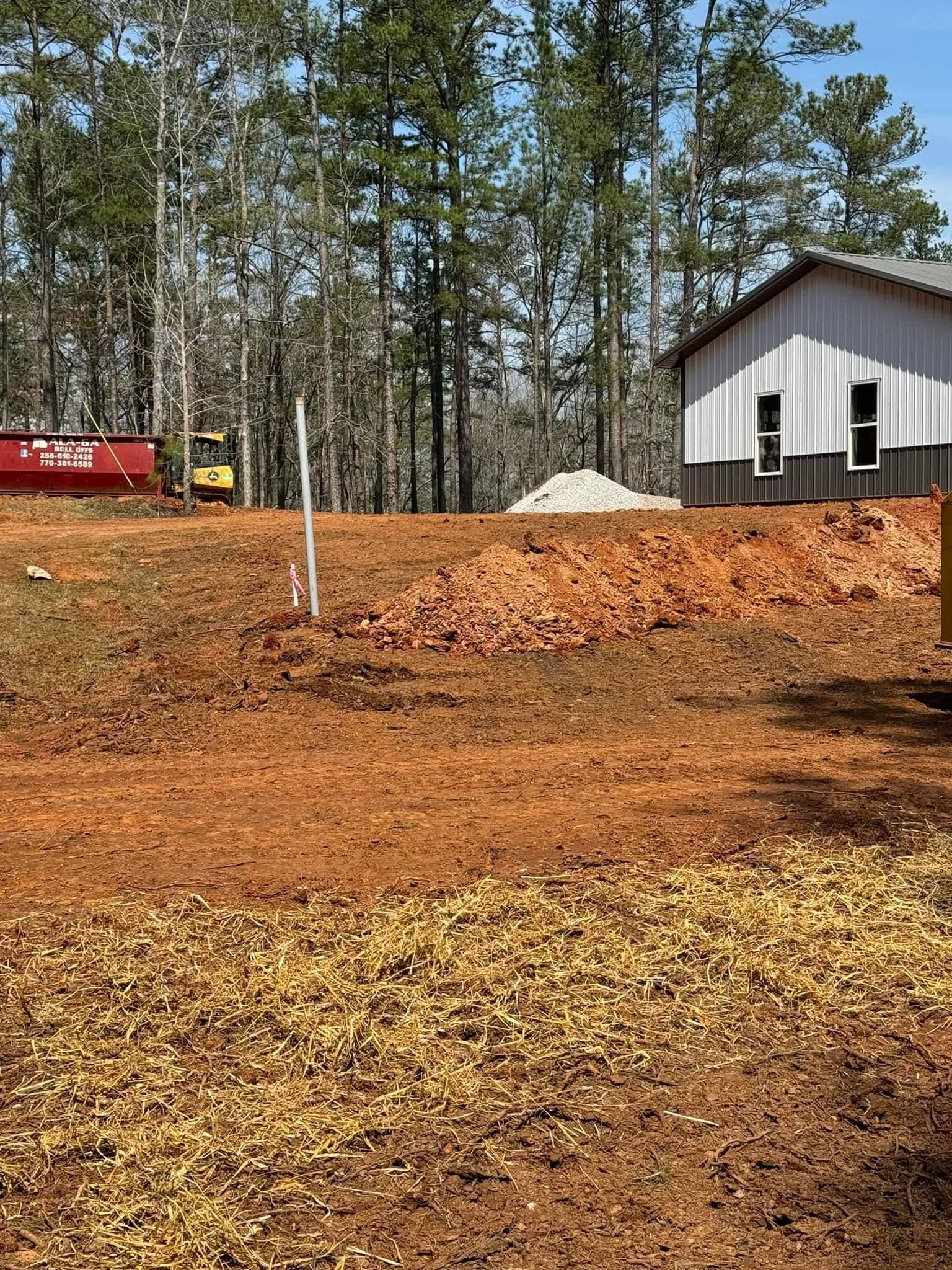 A construction site with a red dumpster, a small excavator, a gravel pile, and a two-toned metal building near trees.
