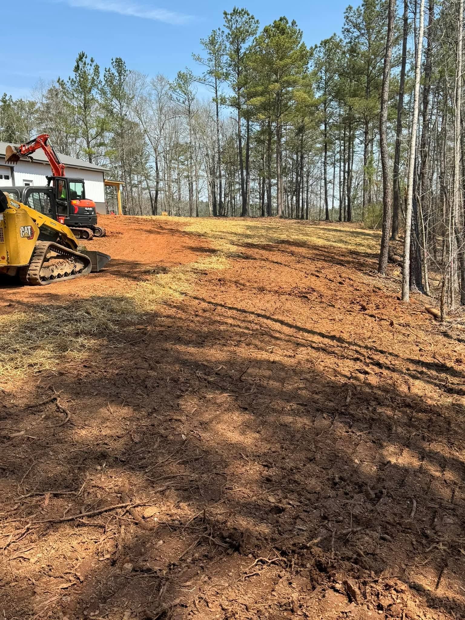 A yellow Caterpillar skid steer clears red dirt on a construction site near a tree-lined forest and a house.