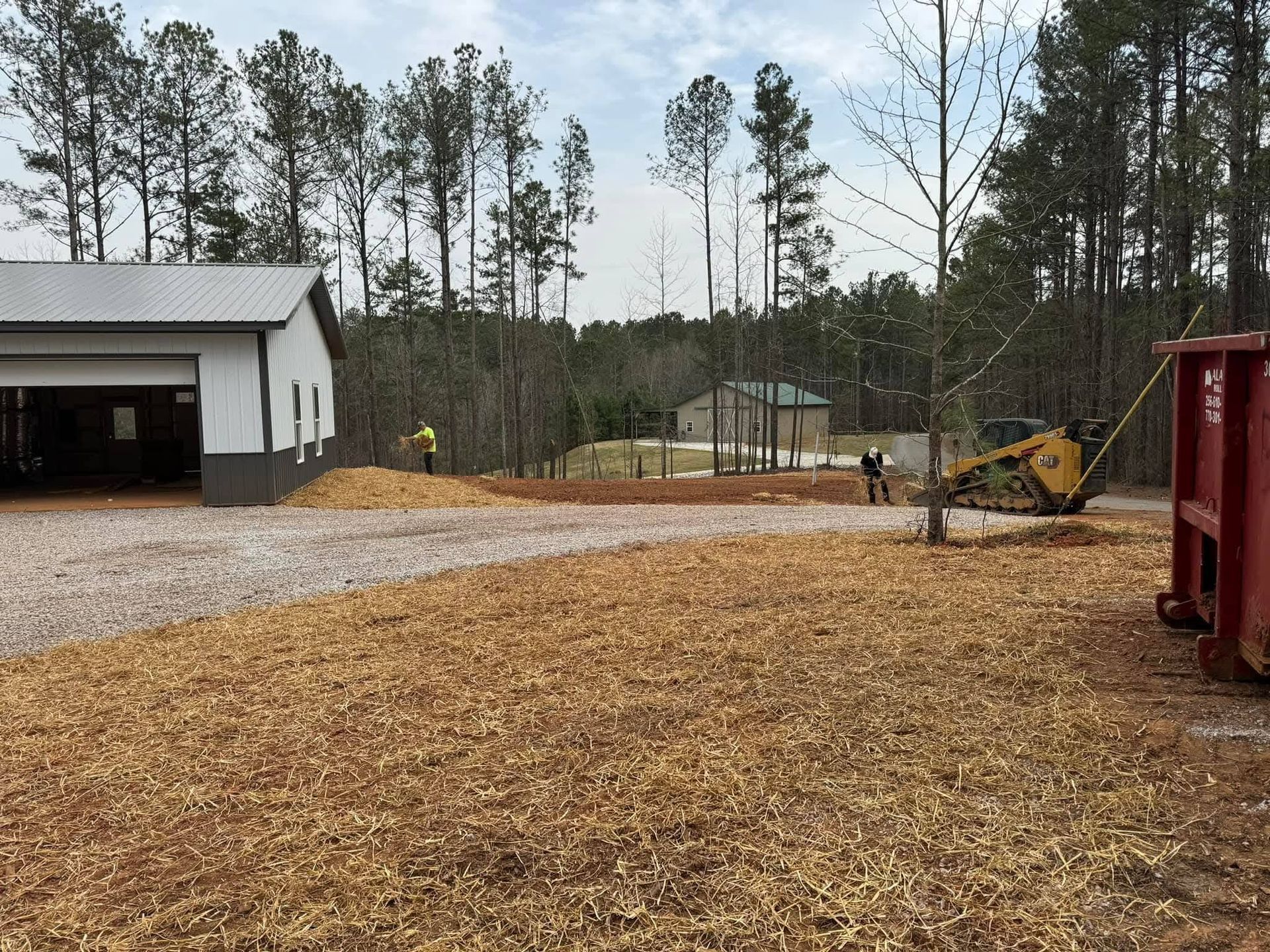 A light-colored building with a large bay door stands near a gravel area, wood chips, a yellow skid steer, and a dumpster.
