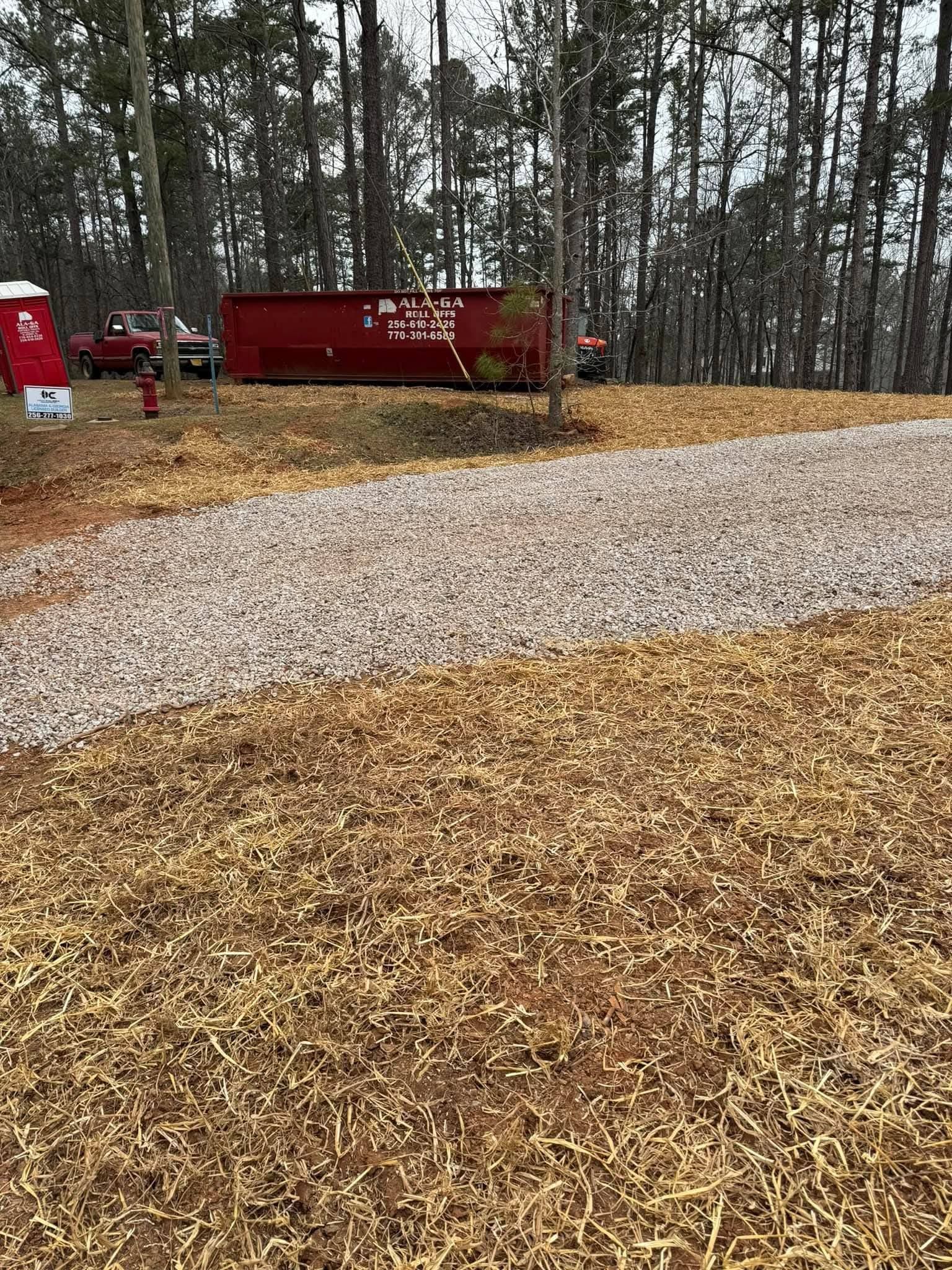 A gravel driveway leads through a wooded area toward a red dumpster and a red truck parked in the background.