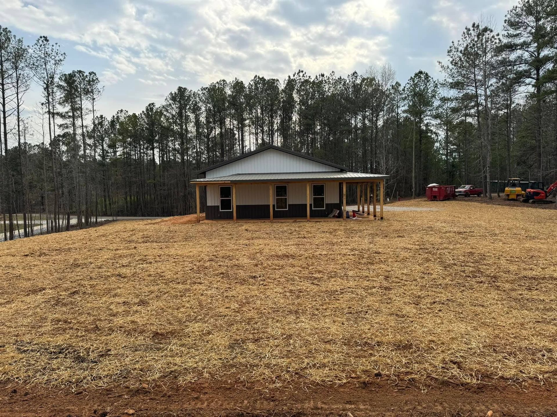 A single-story house with a white exterior and porch sits on a cleared, wood-chipped lot surrounded by a dense forest.