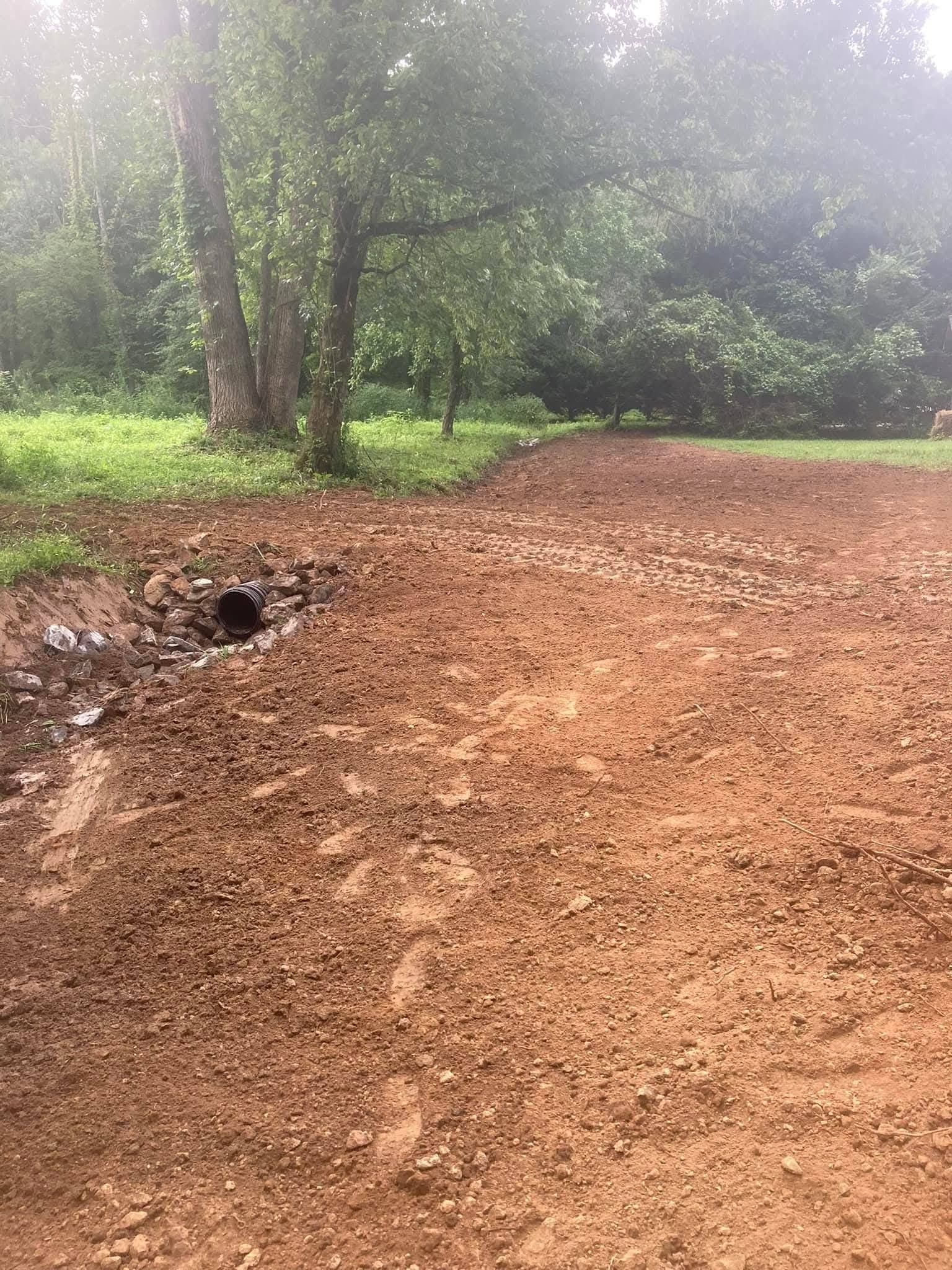 A dirt path leads through a grassy area toward a cluster of trees, with a small drainage pipe visible on the left.